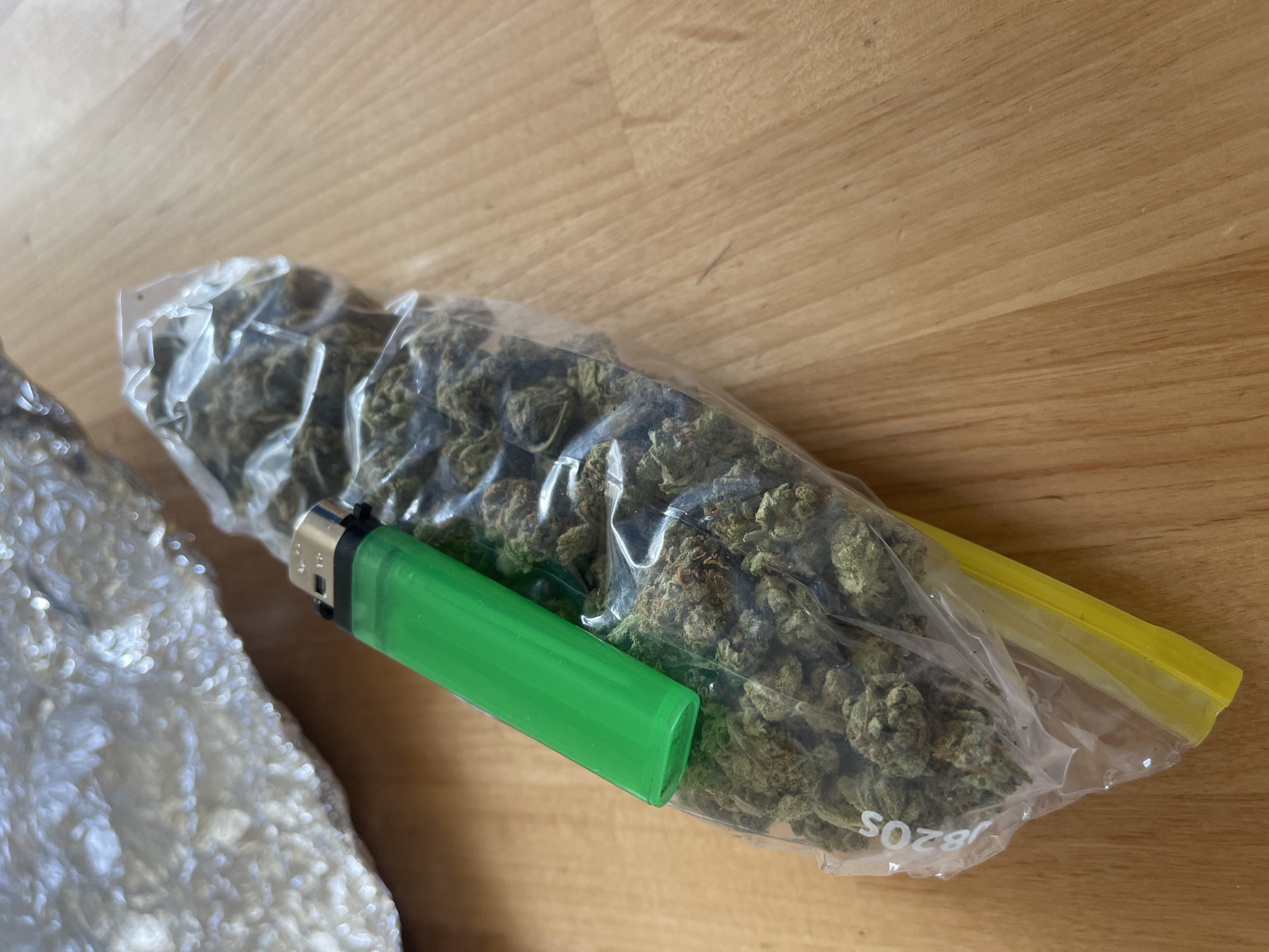 A green lighter rests on a clear plastic bag filled with cannabis. The bag is sealed with a yellow zipper closure. In the background, a crumpled piece of aluminum foil is visible. The objects are placed on a wooden surface.