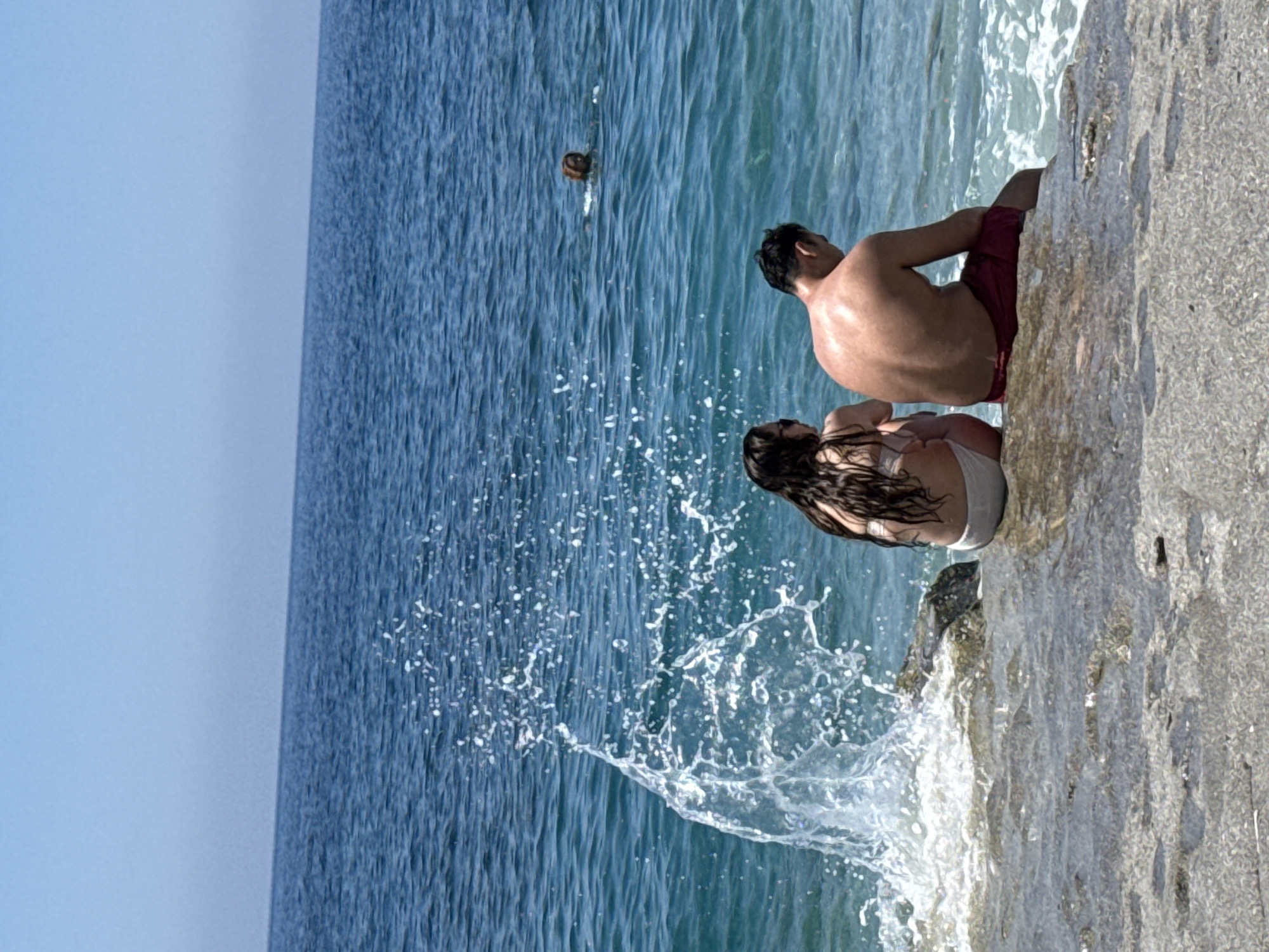 The image shows a man and a woman sitting on a rocky outcrop at the edge of a body of water, likely the sea, as indicated by the waves splashing against the rocks. The man is shirtless and wearing red swim trunks. The woman has long, dark hair and is wearing a white swimsuit. They are positioned close together, possibly interacting or observing the water. A person is swimming in the distance. The sky is clear and blue. The location is the city of Unknown, Unknown.