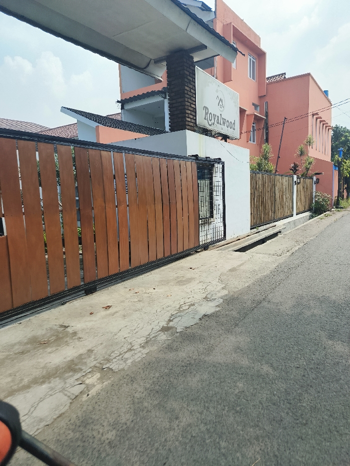 The image shows a gated entrance to a property in Cileunyi, Indonesia, located at coordinates -6.945, 107.726. The gate is made of wooden slats and is closed. A sign above the gate reads "Royalwood." The property is surrounded by a low wall and a small garden area with a few trees. The wall is made of a combination of brick and bamboo. The image is taken from the street, looking towards the gate. The street is paved with asphalt and is relatively empty. There are no people visible in the image. The image is taken from a low angle, looking upwards towards the gate. The sky is visible in the upper left corner of the image. The weather appears to be clear. The image is in focus and well-lit.