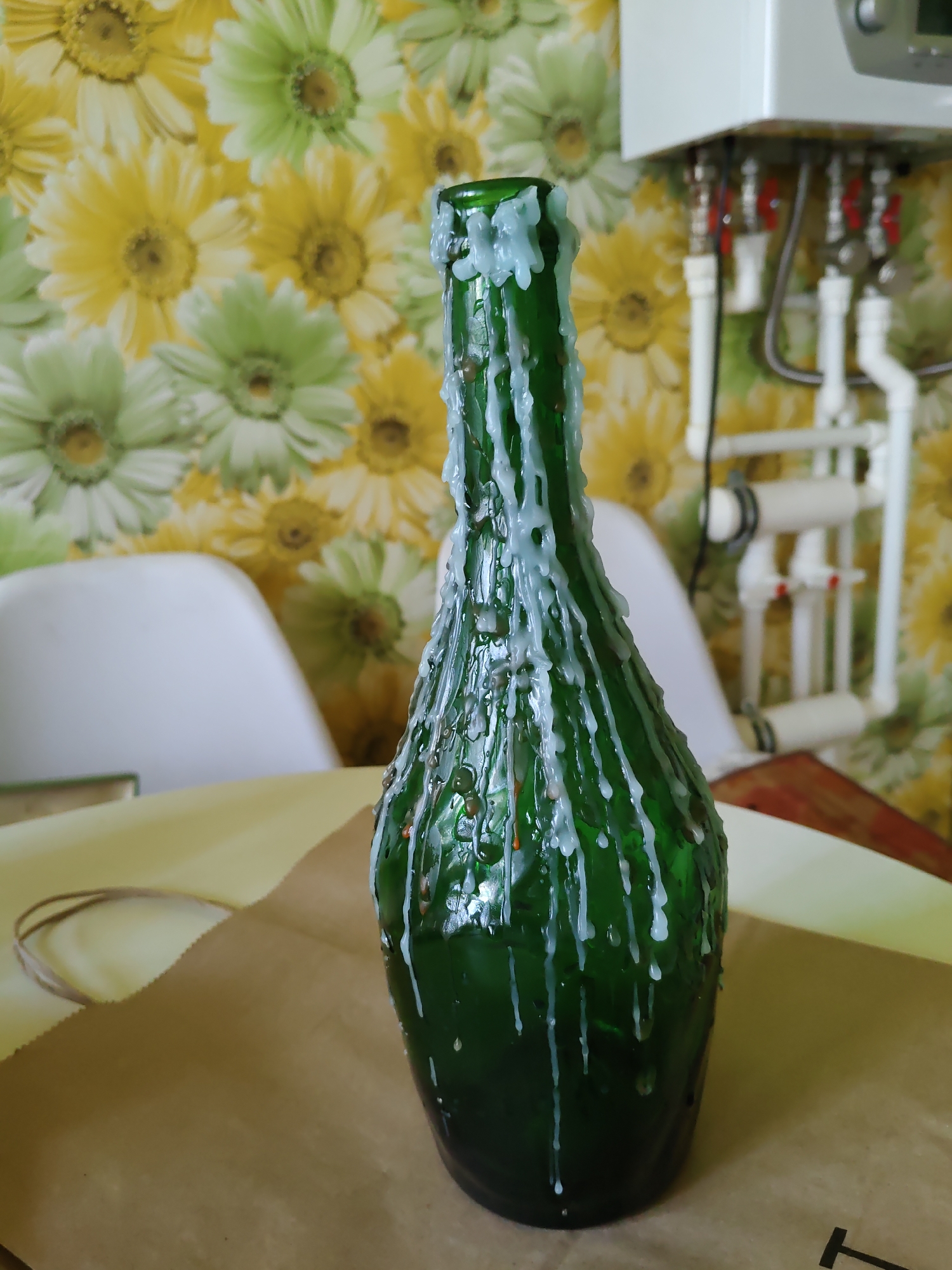 A green glass bottle, covered in dripped white wax, sits on a light yellow surface, partially covered by brown paper. In the background, a wall with a floral pattern of yellow and green flowers is visible, along with a white water heater unit with red-handled valves and pipes. Two white chairs are also partially visible behind the bottle. The scene appears to be indoors. The provided information does not specify a location within Rostov-on-Don, Russia.