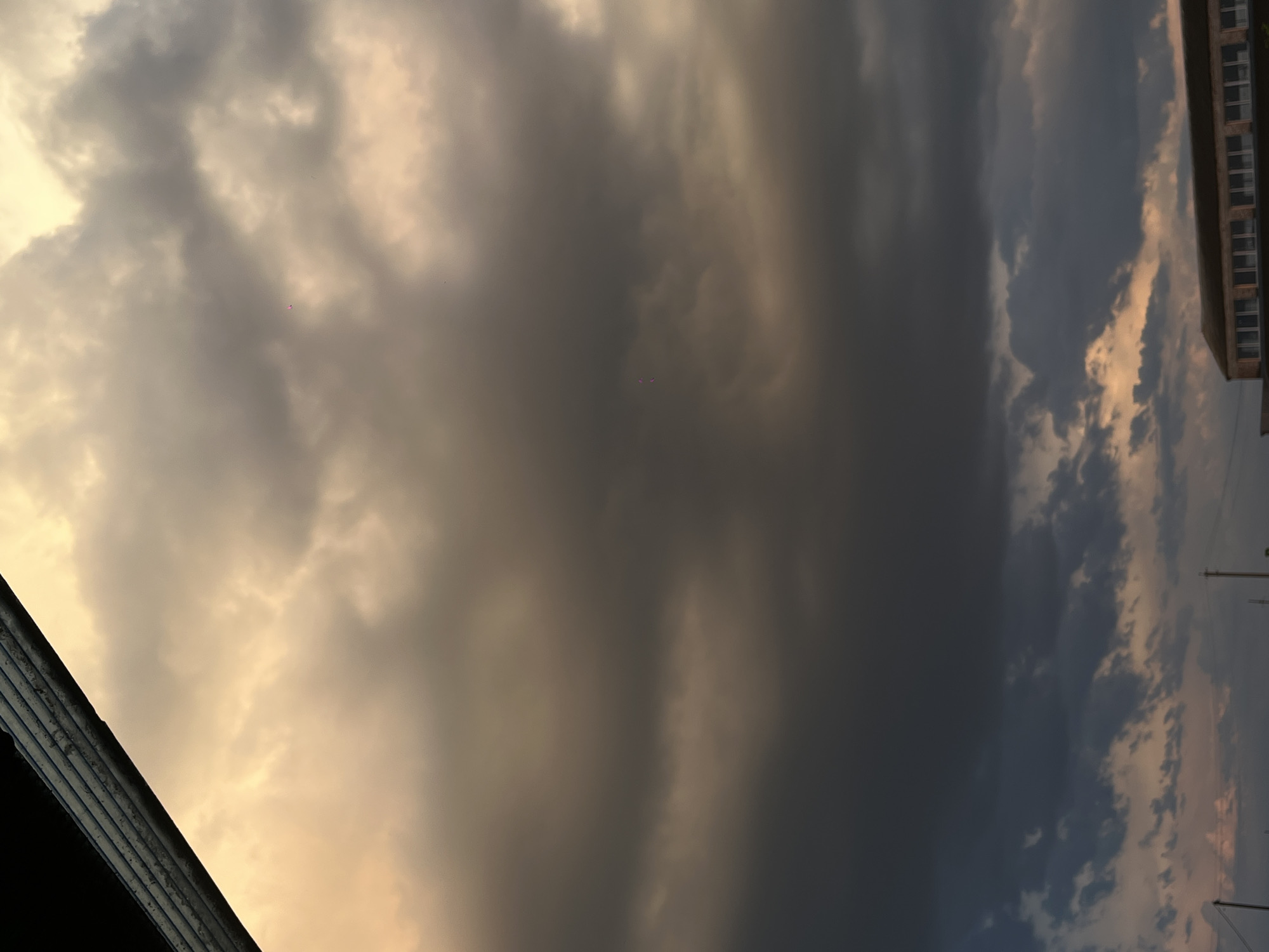 The image shows a cloudy sky over Novopokrovskaya, Russia. The lighting suggests either sunrise or sunset. A building is visible on the right side of the frame. A roof is seen on the bottom left corner. There are power lines on the right side as well.