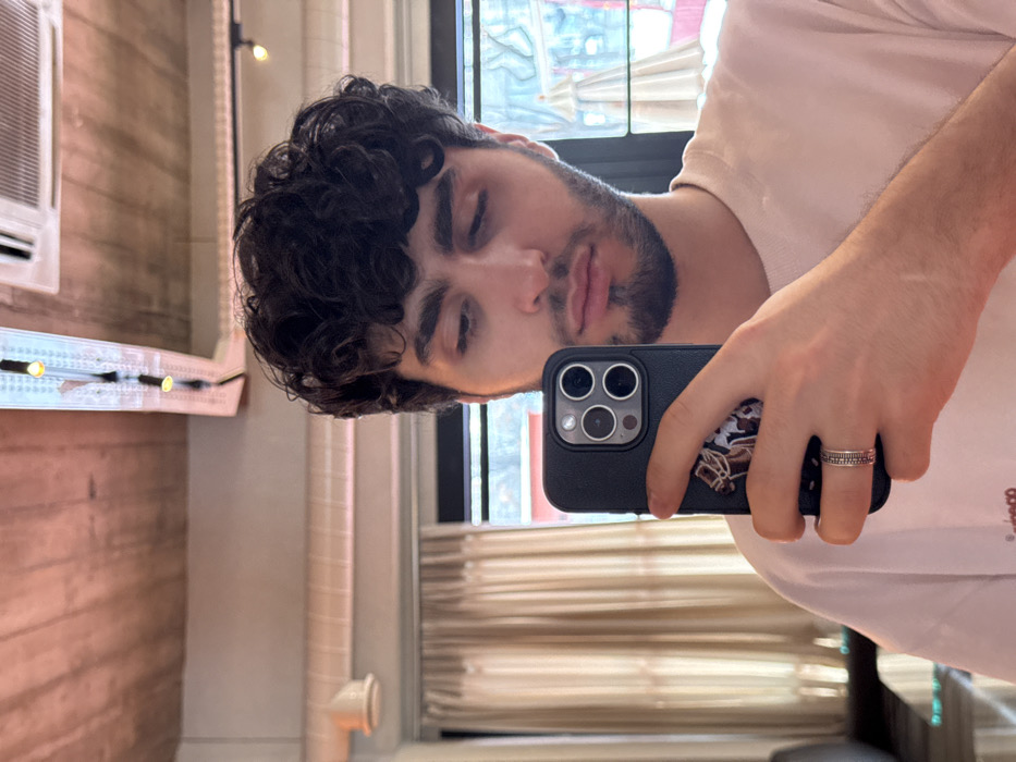 A man with dark curly hair and a short beard takes a selfie in a mirror. He holds a dark smartphone, adorned with a sports team logo, with a silver band on his ring finger. His eyes are closed, and his expression is relaxed. He wears a white t-shirt, revealing the Adidas logo on the sleeve. The reflection shows a bathroom interior with a window, blinds, and a decorative lighting fixture.