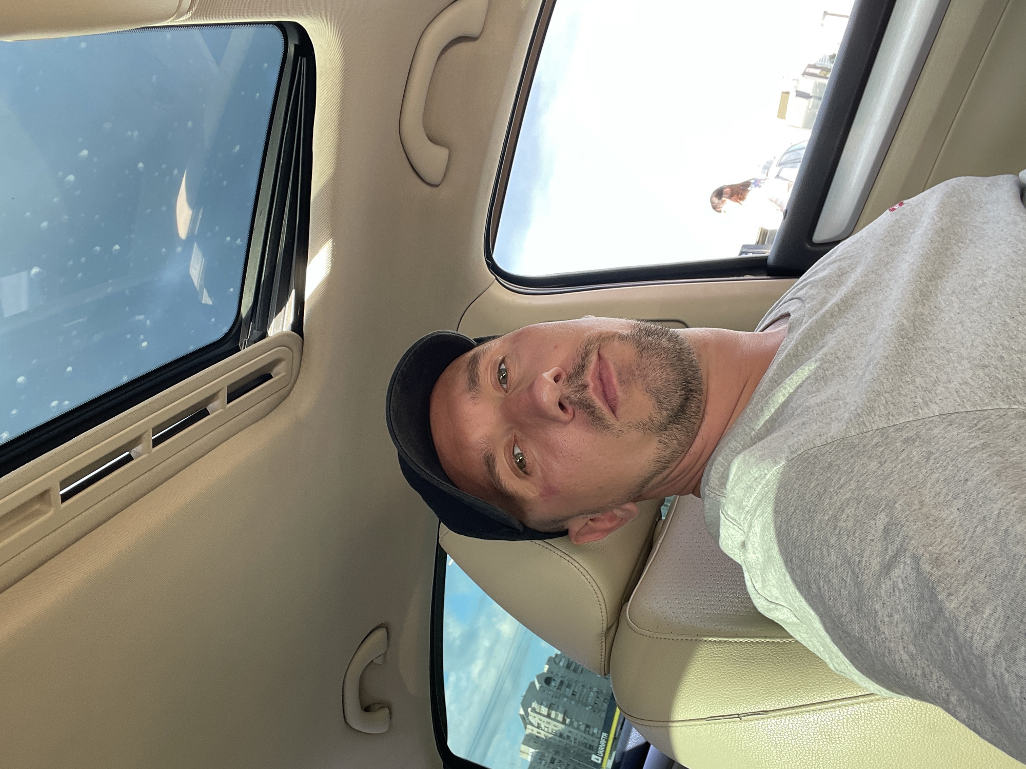 The image shows a man with light skin, short brown hair, a goatee, and green eyes. He is wearing a black baseball cap and a grey t-shirt. He is taking a selfie from inside a car, looking up towards the sunroof. The sunroof is open, and the sky is visible. The interior of the car is light beige.
In the background, through the open sunroof, there are some buildings and a clear blue sky with some clouds. There are also some reflections on the sunroof glass, including what appears to be a person. The time of day appears to be daytime, likely sunny. The scene is set inside a vehicle, possibly a car, with a view of the outside through the sunroof.
There is no visible text in the image. The man's expression is neutral. The image is taken from a low angle, looking up.