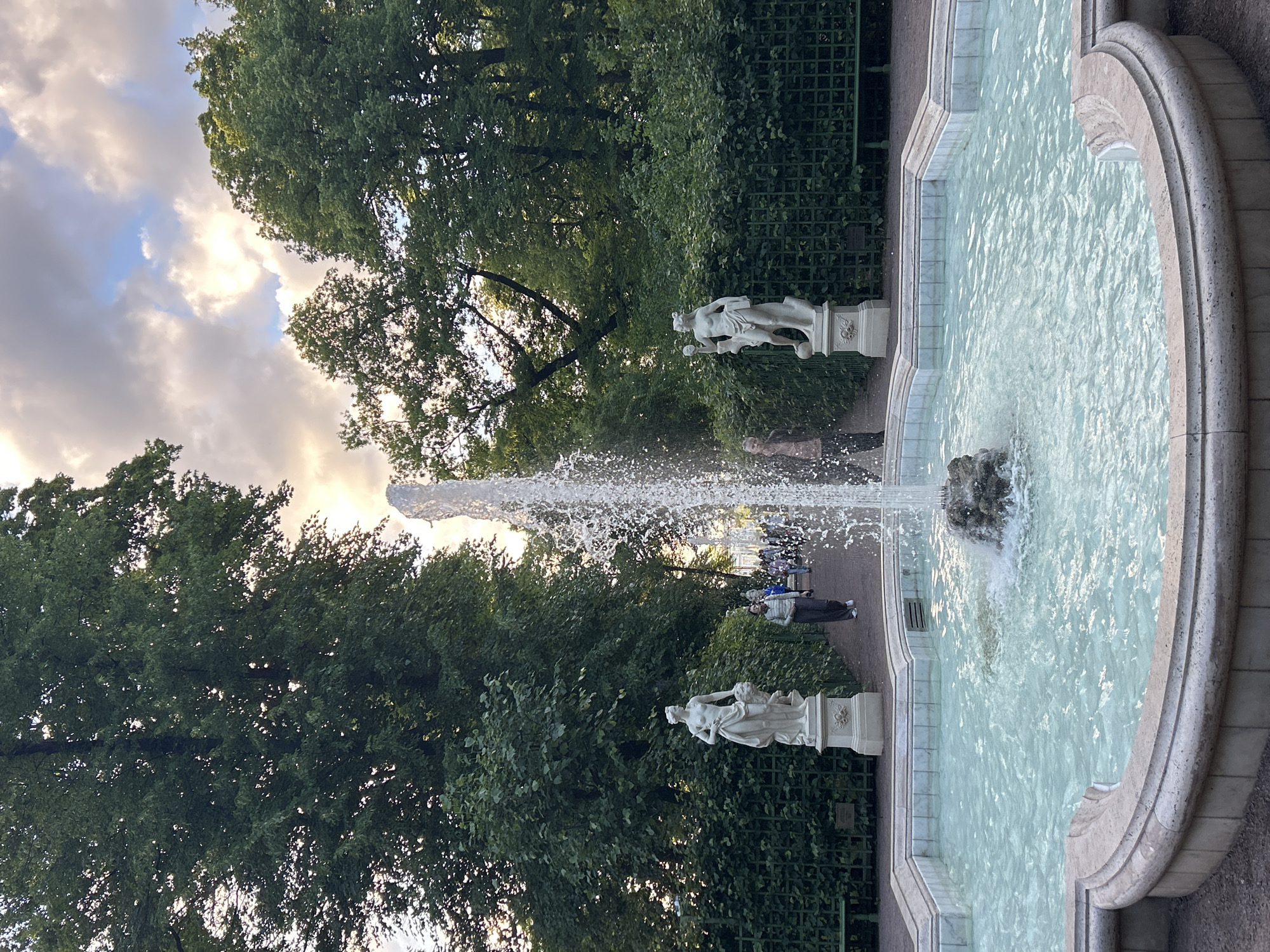 The image shows a fountain in a park. The fountain has a white stone basin and water is spraying from a rock formation in the center. On either side of the fountain, there are white stone statues of women. In the background, there are people walking on a path, and large green trees. The sky is partly cloudy with some sunlight breaking through.