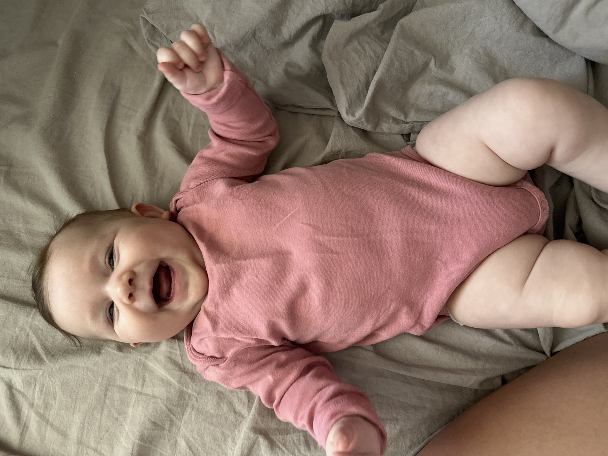 A baby wearing a pink bodysuit lies on a gray sheet, smiling widely with an open mouth. The baby's left arm is raised with a clenched fist. The baby has fair skin and dark hair. The location is unspecified and the city of Chaikovsky, Russia, is not discernible in the image.
