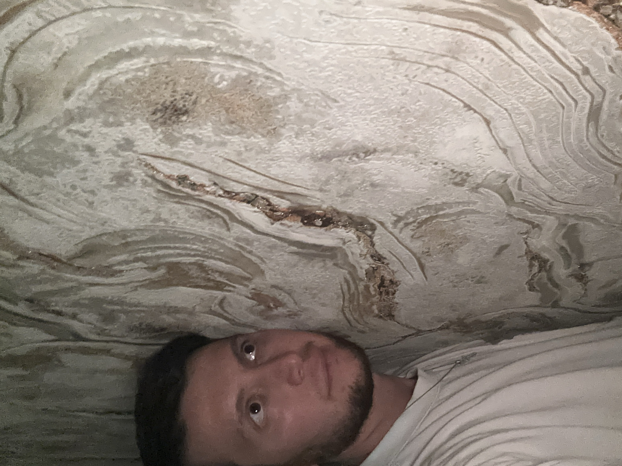 A man with dark hair and a beard is leaning against a textured wall. He is wearing a light-colored t-shirt. The wall has a rough, layered texture with swirling patterns in shades of white, beige, and brown. The man is looking upwards and to the right, with his eyes open. The scene appears to be indoors, possibly in a cave or a building with decorative wall treatment. The location is identified as Mirny, Russia.