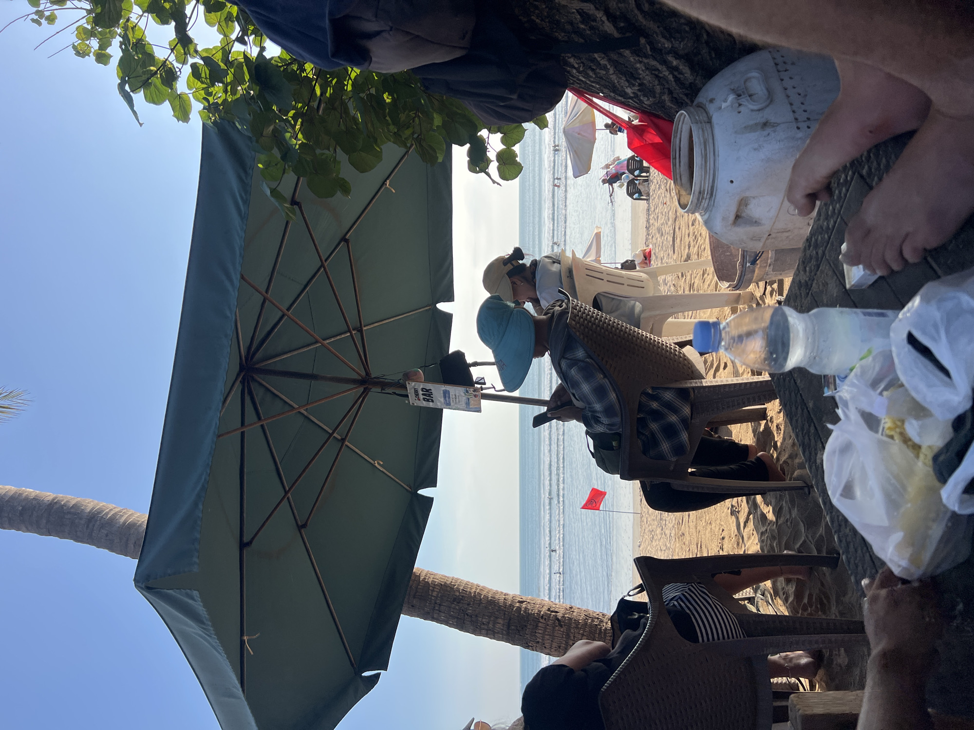 The image depicts a beach scene in Kuta, Indonesia, under a large, dark green umbrella. In the foreground, a table is laden with food items in plastic bags and a bottle of water. A person's legs and hands are visible, suggesting they are seated at the table. Behind the table, several individuals are seated on chairs, facing the ocean. One person, wearing a plaid shirt and a blue hat, is looking at their phone. Another person, wearing a lighter colored hat, is looking towards the sea. The beach itself is sandy, with a red flag planted in the sand. The ocean is visible in the background, with a few people in the water. A palm tree trunk is to the left of the frame, and foliage is on the right. A sign for "CHERRY BAR" hangs from the umbrella. The overall atmosphere suggests a relaxed day at the beach.