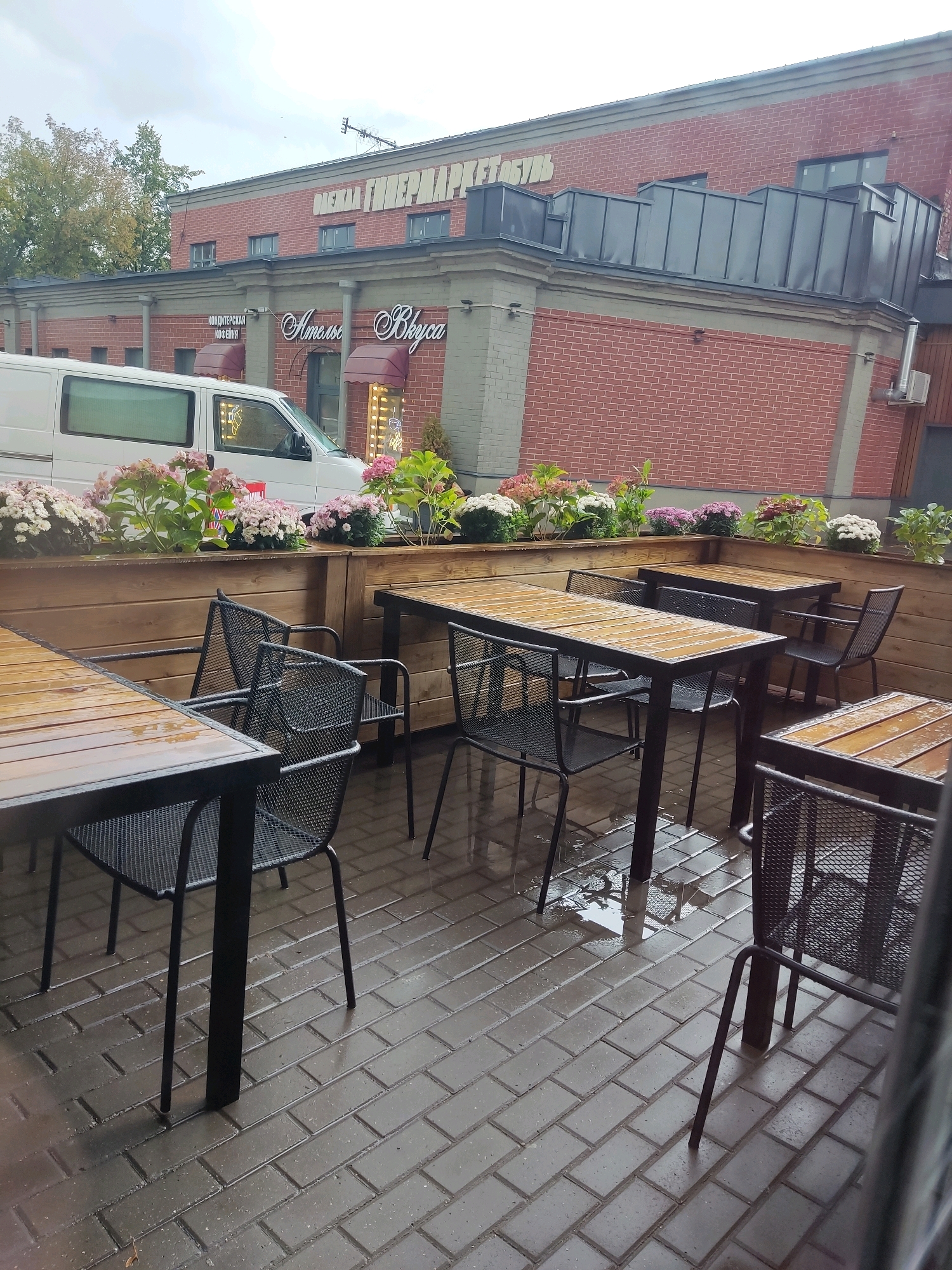 An outdoor seating area of a cafe is depicted on a cloudy day. The ground is wet and reflective, indicating recent rain. Several wooden tables with black metal frames are set with black metal mesh chairs. In the background, a white van is parked next to a brick building with red brick walls and gray accents. Above the van, signage on the building reads "ОЛЕКСА ГИПЕРМАРКЕТ ОБУВЬ" and "Алые Вкусы". The scene appears to be located in Noginsk, Russia, as suggested by the signage which is in Russian and the overall architectural style. Flowers in planters line the wooden partition separating the seating area from the building.