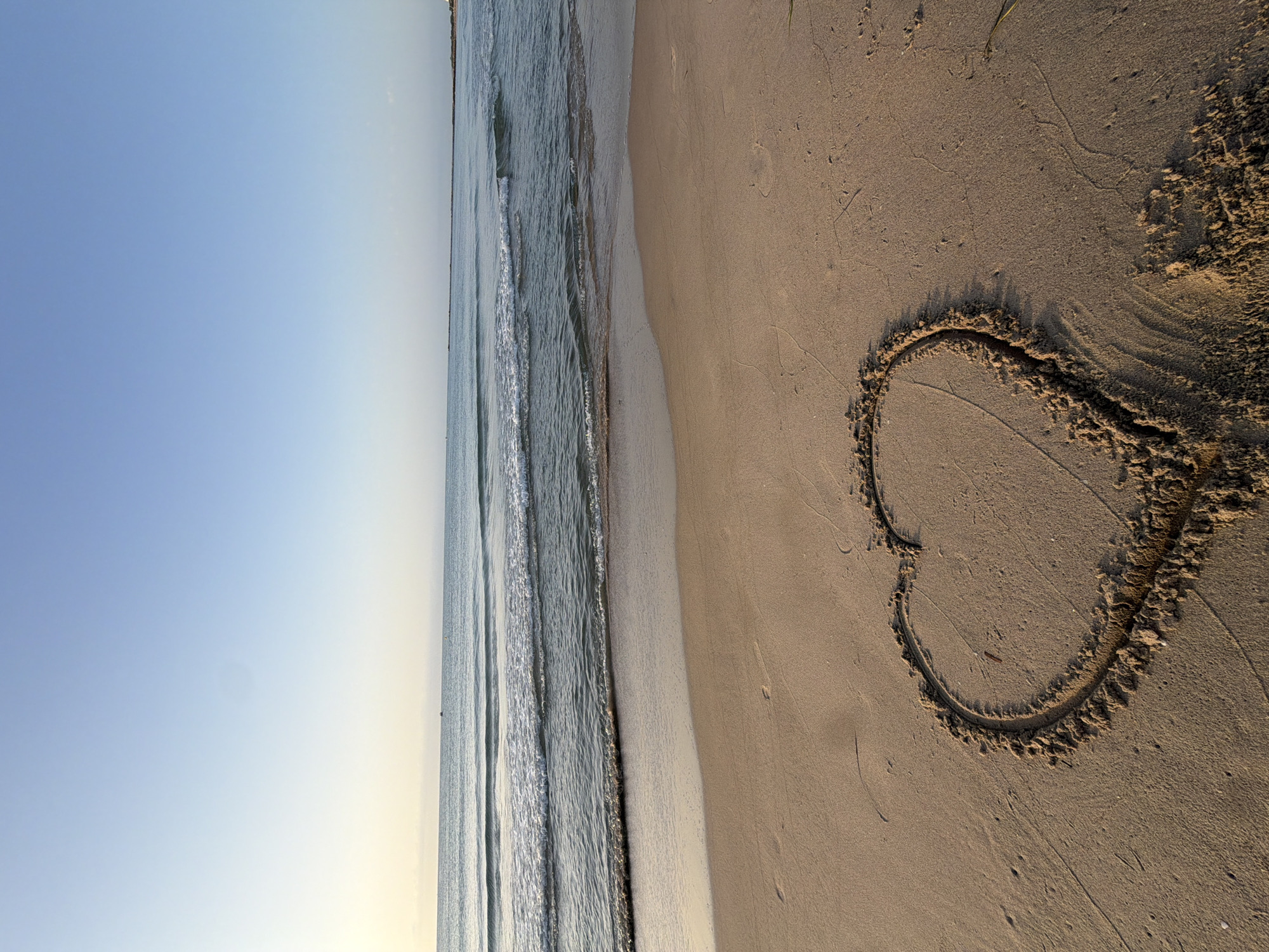 A heart shape is drawn in the sand on a beach. The beach is sandy and appears to be at the sea. The sea is calm with small waves. The sky is clear and blue, transitioning to a lighter shade near the horizon. The scene is likely in El Campello, Spain, given the context.