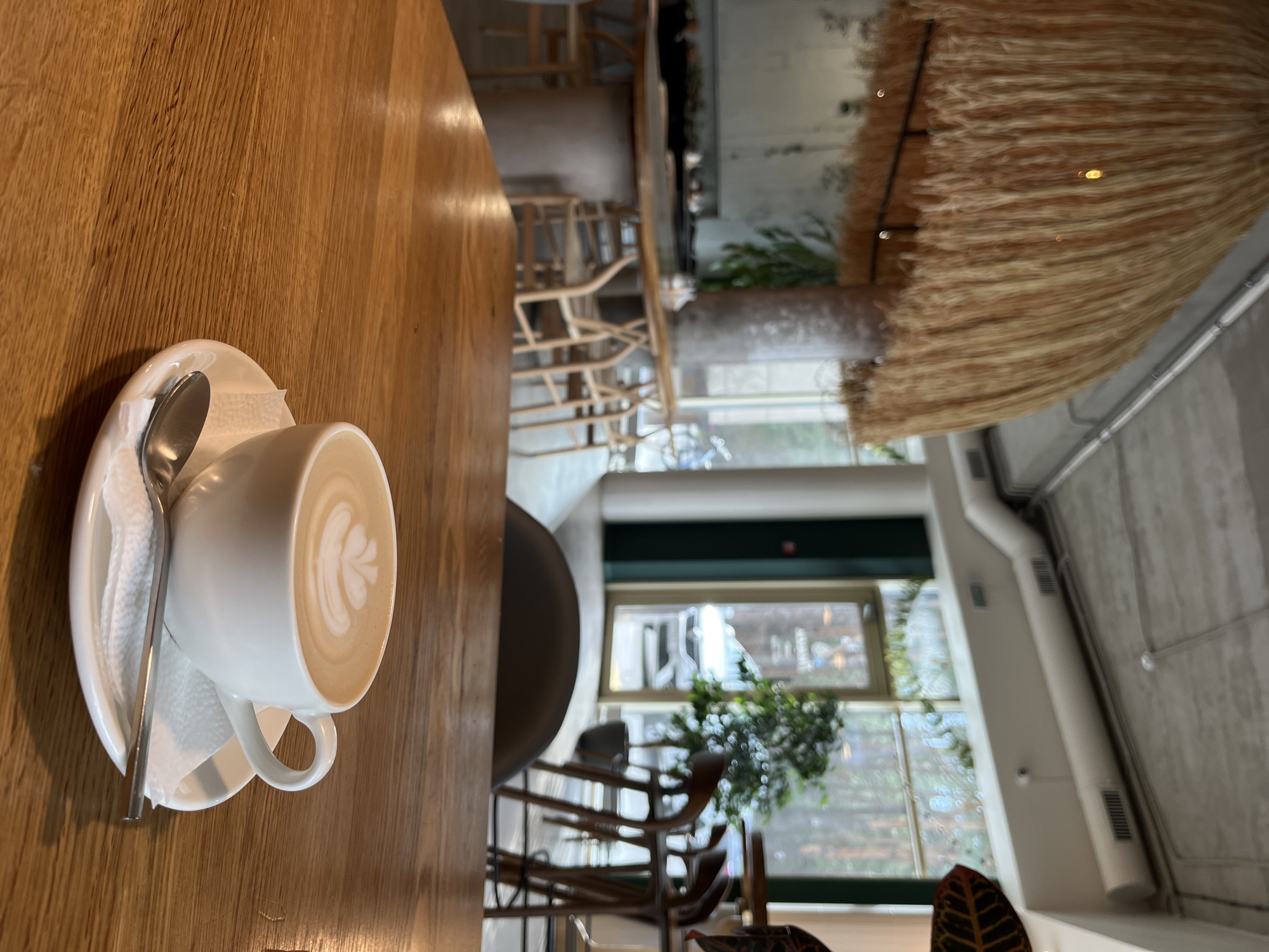 A high-angle shot shows a cup of latte art on a wooden table in a cafe in Hrodna, Belarus. The latte is in a white ceramic cup on a white saucer, with a silver spoon. A folded white paper napkin is underneath the spoon and cup. The latte art is a leafy floral design. The table is made of light-colored wood. In the background, there are chairs and a window. A large, round, straw-like decorative object is suspended from the ceiling. Through the window, the surrounding buildings are visible.