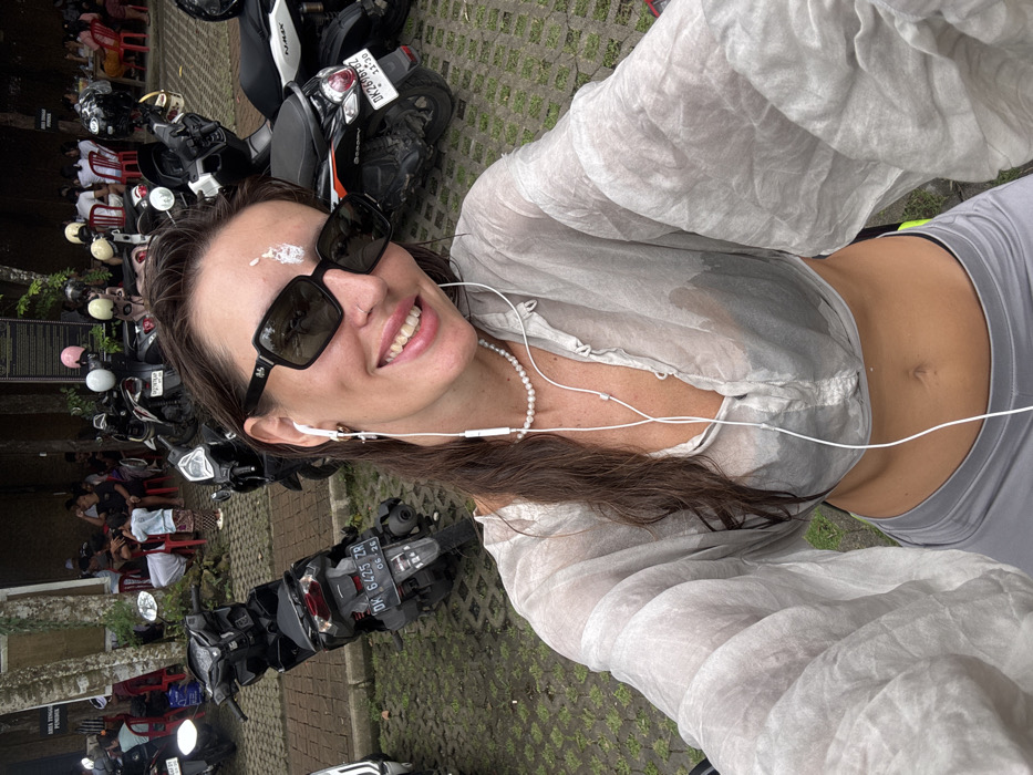 A woman with long brown hair takes a selfie, wearing sunglasses and earbuds, with a white substance on her forehead. She has on a sheer, long-sleeved shirt, a pearl necklace, and gray leggings. Parked motorbikes fill the background, along with a row of people seated in red chairs. The ground is paved with patterned bricks. The license plate of one motorbike is partially visible.
