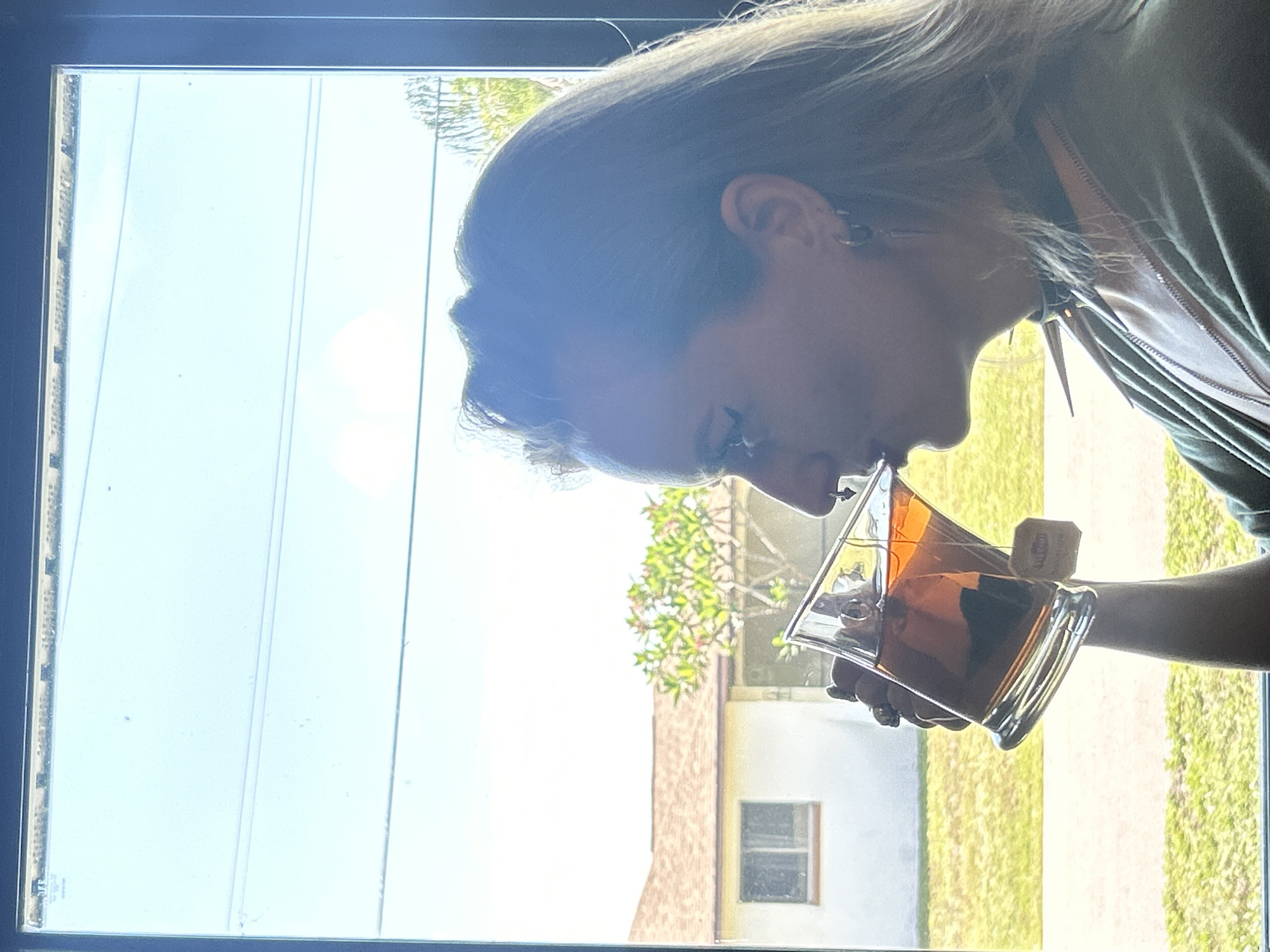 A side profile of a woman drinking tea from a large glass in front of a window. The woman is wearing a green shirt, a black choker, and hoop earrings. A tea bag is visible in the glass, with the tag hanging over the rim. Outside the window, a white house with a brown roof and a green lawn are visible. The sky is bright blue with some wispy clouds. The scene appears to be in a residential area in Cape Coral, Florida.