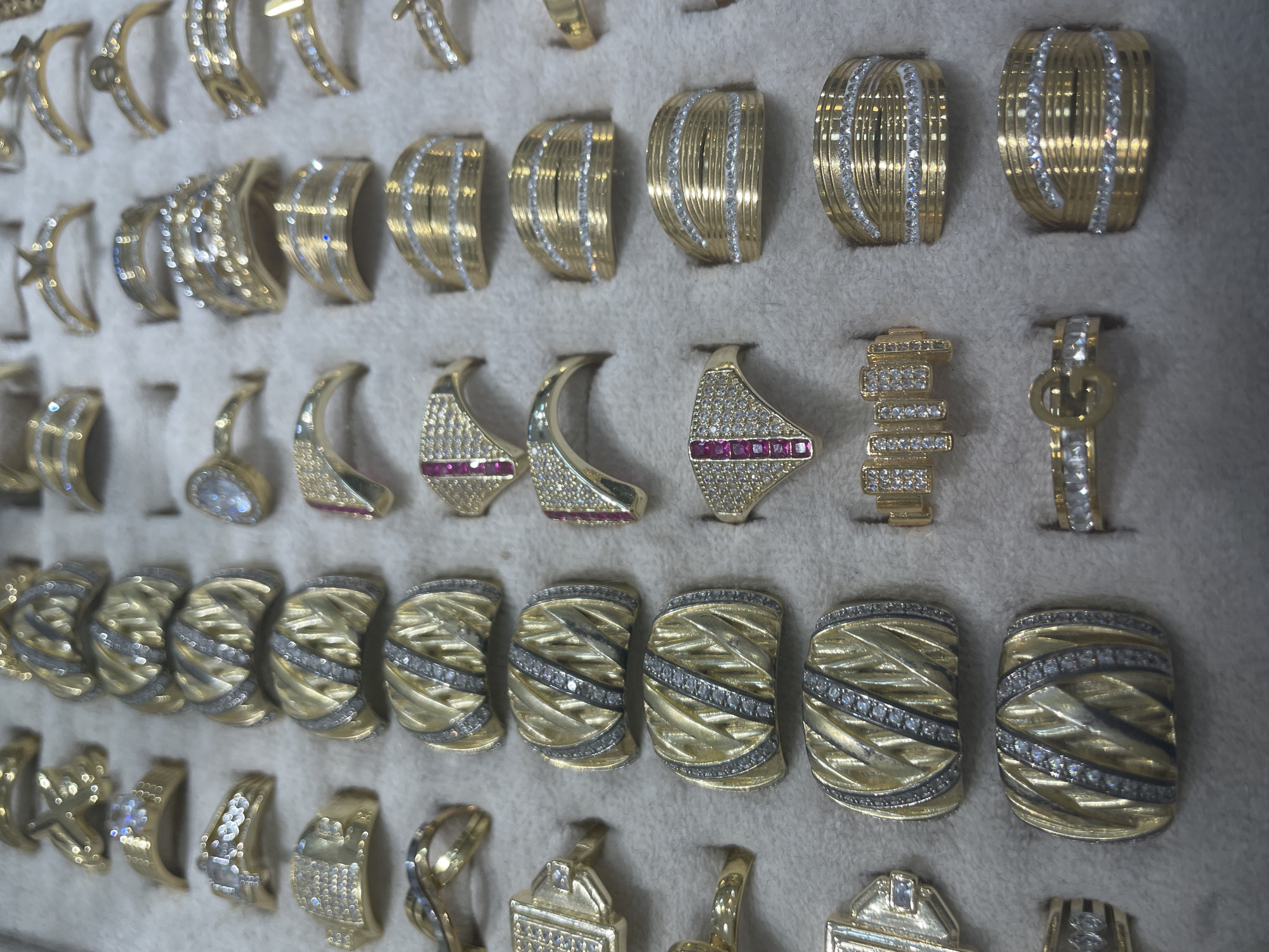 This media file is a close-up, eye-level shot of a collection of gold rings displayed on a grey fabric surface, likely in a jewelry store or personal collection. The rings are arranged in rows and columns, with some individual pieces and some pairs. They vary in design, with some featuring intricate patterns, while others are adorned with small, clear gemstones that appear to be diamonds or cubic zirconia, and a few have red stones, possibly rubies. The gold appears to be yellow gold, and the lighting highlights the shine and detail of each piece. There are no people visible in the image, nor any specific time of day, weather, or strong emotional cues. The setting is generic, focusing solely on the jewelry. There is no visible text in the image. The context provided indicates the location is Yunus Emre, Turkey, which might suggest a particular style or origin of the jewelry, but this is not evident from the visual information alone.