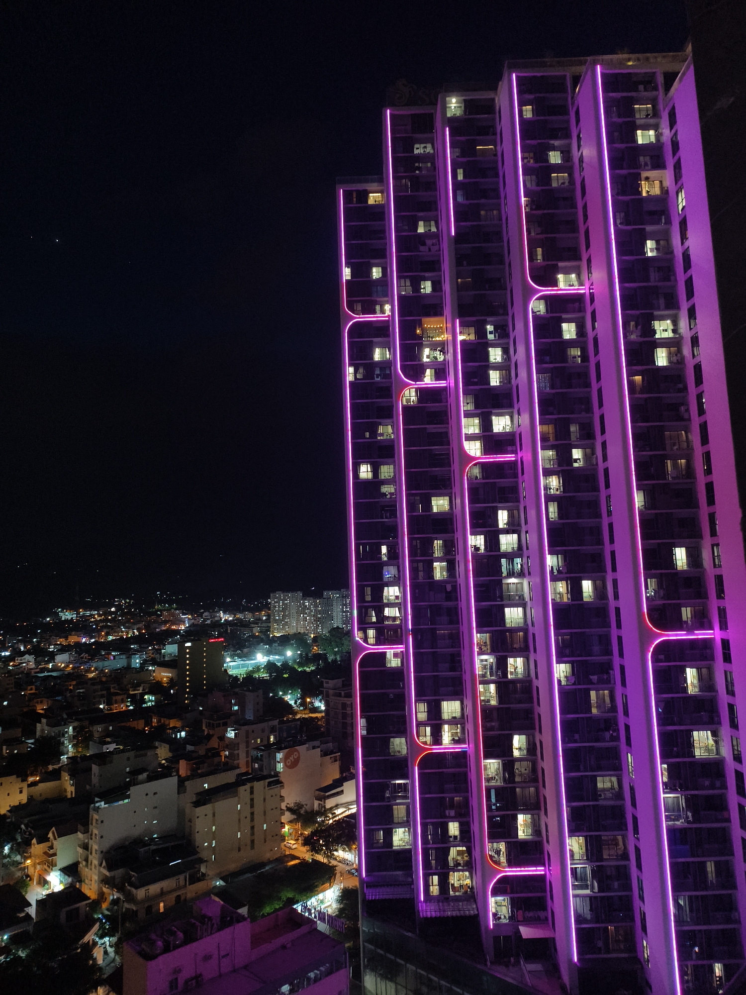 This is an aerial, nighttime photograph of a city with a focus on a tall, modern, white skyscraper illuminated by vibrant pink LED lights. The skyscraper features a unique design with vertical columns and horizontal bands, accented by flowing, curved lines of light that outline its architectural features. Numerous windows are visible on the skyscraper, with some illuminated from within, suggesting occupancy.
To the left of the skyscraper, a sprawling cityscape unfolds under the dark sky, dotted with numerous buildings of varying heights and architectural styles. The city is lit by a combination of streetlights, building lights, and the glow from distant structures. Several smaller buildings are visible in the foreground, appearing to be residential or commercial structures. A sign with the red "OYO" logo is discernible on one of the mid-sized buildings in the mid-ground. In the far distance, more densely packed, brightly lit buildings suggest a more developed urban area. The overall atmosphere is one of urban vibrancy and nighttime activity.