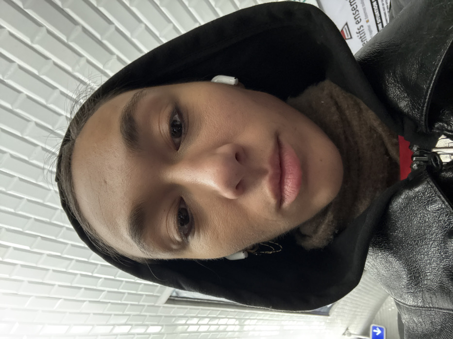 A young adult, appearing female, is depicted in a close-up, downward-facing selfie. The individual has a neutral expression, with lips slightly parted and eyes directed towards the camera. They wear a black hooded jacket over a brown turtleneck, with a glimpse of red fabric visible beneath. White wireless earbuds are present in both ears, and a small, gold-colored chain earring is visible on the subject's left earlobe. The background features a distinctive white tiled ceiling composed of rectangular, beveled tiles arranged in a staggered pattern. To the lower left, a blue directional sign with a white arrow pointing left is partially visible next to a vertical glass or reflective surface. To the lower right, a white sign or poster with black French text is partially visible, displaying fragments such as "entifs ensemb" and "situation." This environment, including the specific tile pattern, signage, and language, identifies the location as a station within the Paris Metro system in Paris, France.
