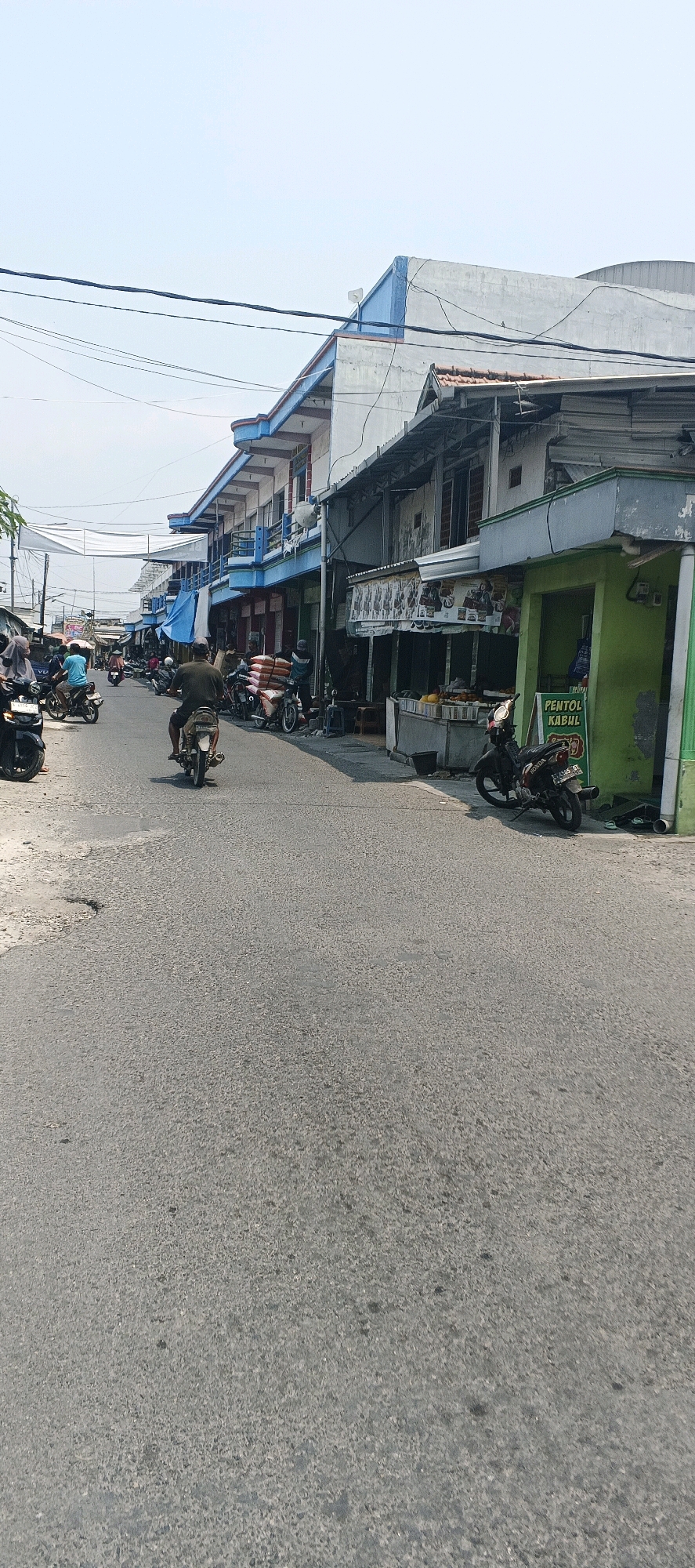 This image depicts a street scene in Mayong, Indonesia, likely during daylight hours, with clear skies overhead. The street is paved with asphalt and shows some wear, including visible potholes. On either side of the street are multi-story buildings that appear to be shops or residences, with some featuring blue and white facades.
Several people are present in the scene, mostly on or around motorcycles. In the foreground, a person is riding a motorcycle away from the camera. Further down the street, other individuals are seen on motorcycles, some parked and others in motion. Some buildings have awnings or stalls extending onto the sidewalk area, suggesting commercial activity.
One prominent structure on the right side of the street is a green building with an open front, possibly a shop. A sign on this building reads "PENTOL KABUL". There is a food stall with fruits displayed in front of it. Another stall further down shows various food items or products under a banner. Stacks of goods, possibly sacks of rice, are seen loaded onto a motorcycle. Electrical wires are strung across the street and between buildings. The overall impression is of a busy, somewhat modest urban commercial area.