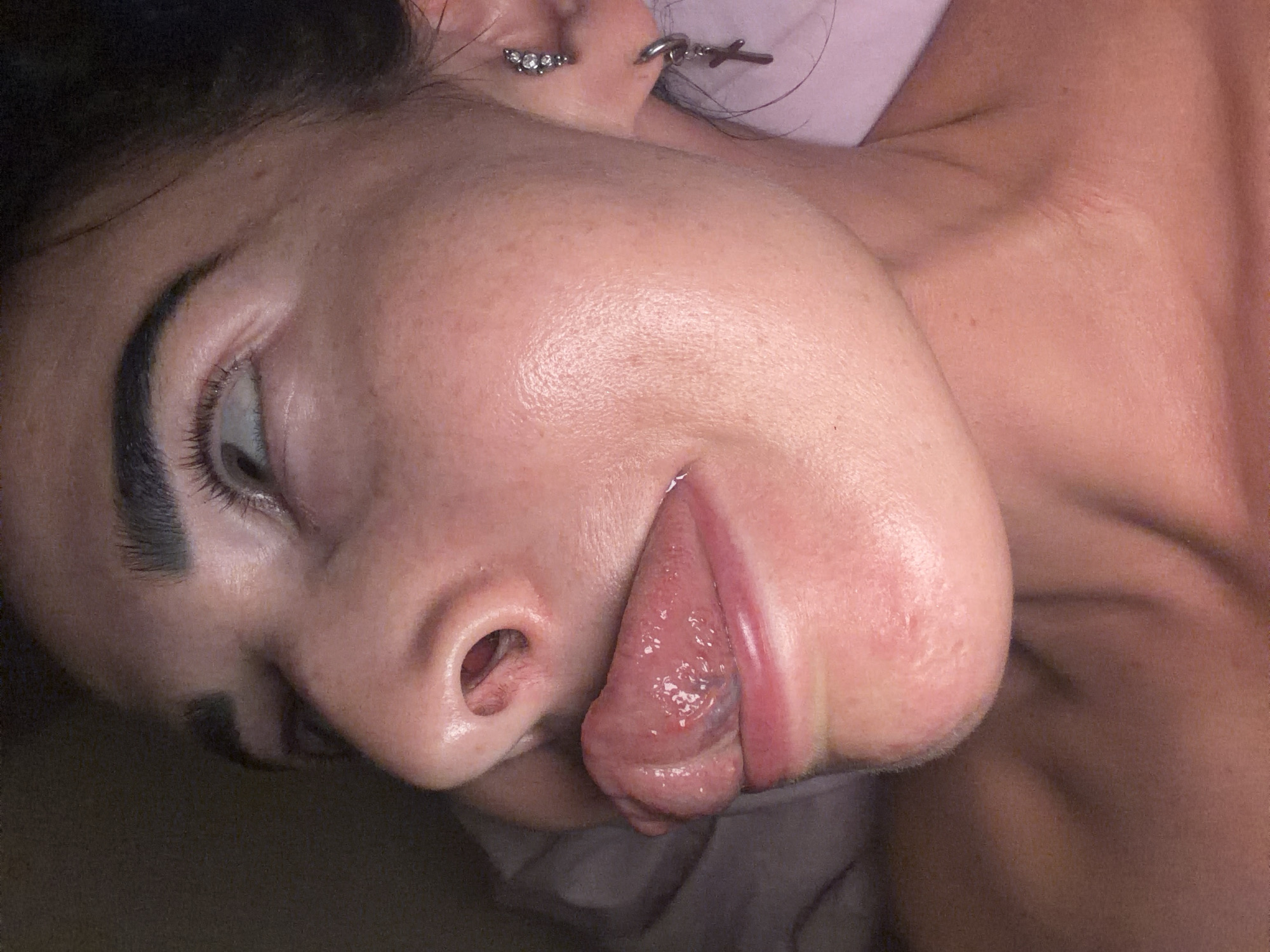This media file presents a close-up portrait of a person, likely female, captured from the neck up, with their head tilted to the side. The subject is seen in profile, lying down, with dark hair framing their face. They possess distinct, dark eyebrows and eyelashes, and their left eye is partially open, gazing upwards.
The central activity captured is the person extending their tongue out to the side of their mouth, creating a playful or expressive facial gesture. The texture and prominent veins of the tongue are clearly visible in detail. Two earrings adorn the visible left ear: one is a cluster of small, sparkling stones, and the other is a silver-toned hoop featuring a dangling cross charm.
The person's skin appears smooth with a subtle sheen, suggesting either natural oils or moisturizer, and a few faint freckles or blemishes are discernible. The setting appears to be indoors, possibly on a bed or soft surface, indicated by light-colored fabric behind the head and neck. The lighting is soft and ambient, making the specific time of day indeterminate. The overall mood conveyed is one of informality and lightheartedness.
There is no visible text in the image. While the provided context indicates Saint Petersburg, Russia, there are no specific visual cues within the image itself to confirm this location.