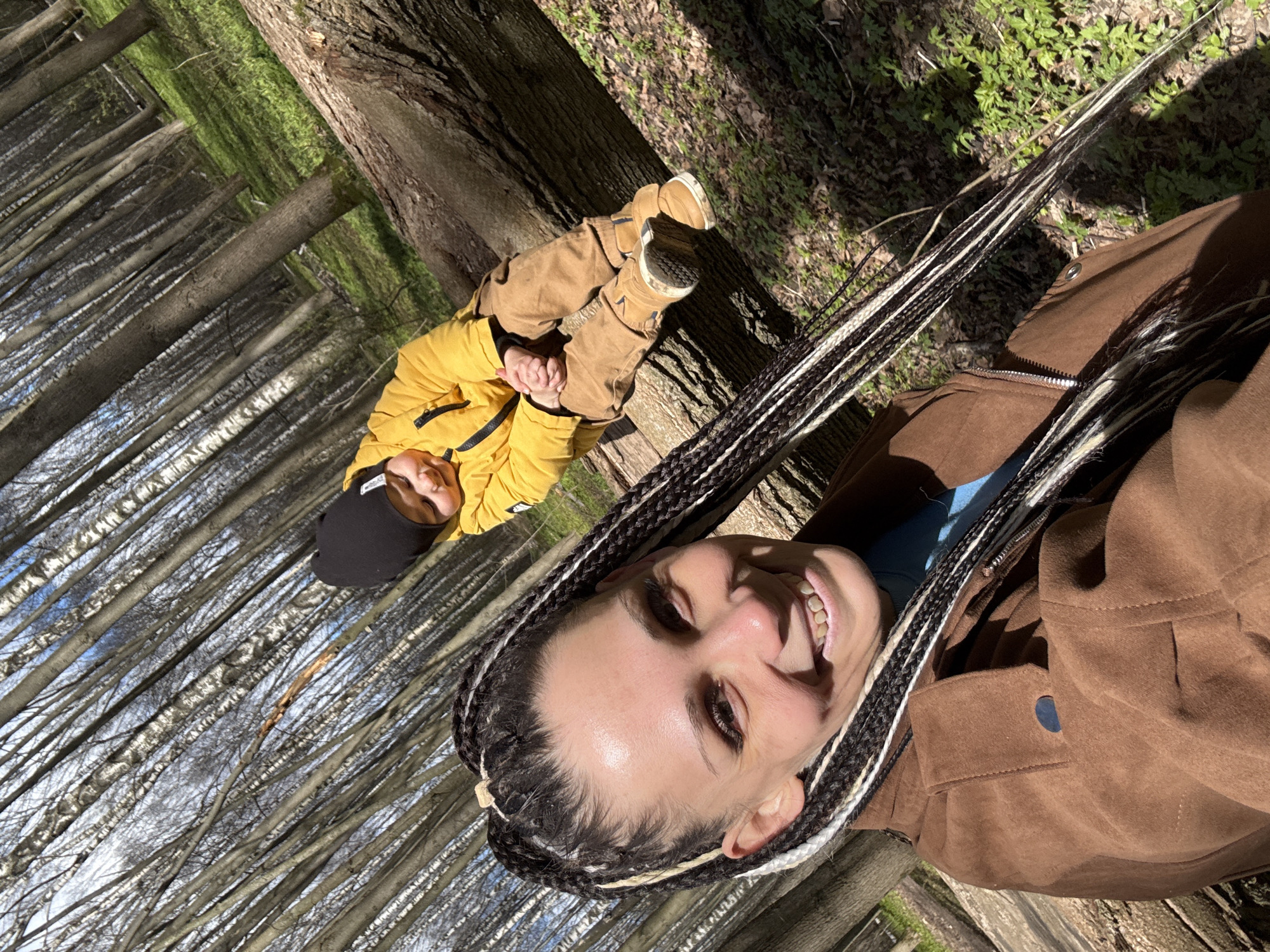 A medium shot shows a woman and a child, both in autumn attire, in a forest. The woman, situated in the foreground on the right, is wearing a brown suede jacket and has long, braided hair with interspersed white strands. She is smiling at the camera. To her left, slightly blurred in the background, a child clad in a yellow puffy jacket, brown pants, and brown shoes, lies on a tree trunk. The trees in the forest are bare, with slender trunks forming parallel lines. Sunlight filters through the trees, creating a bright, natural atmosphere. The location is in the city of Unknown, Unknown.