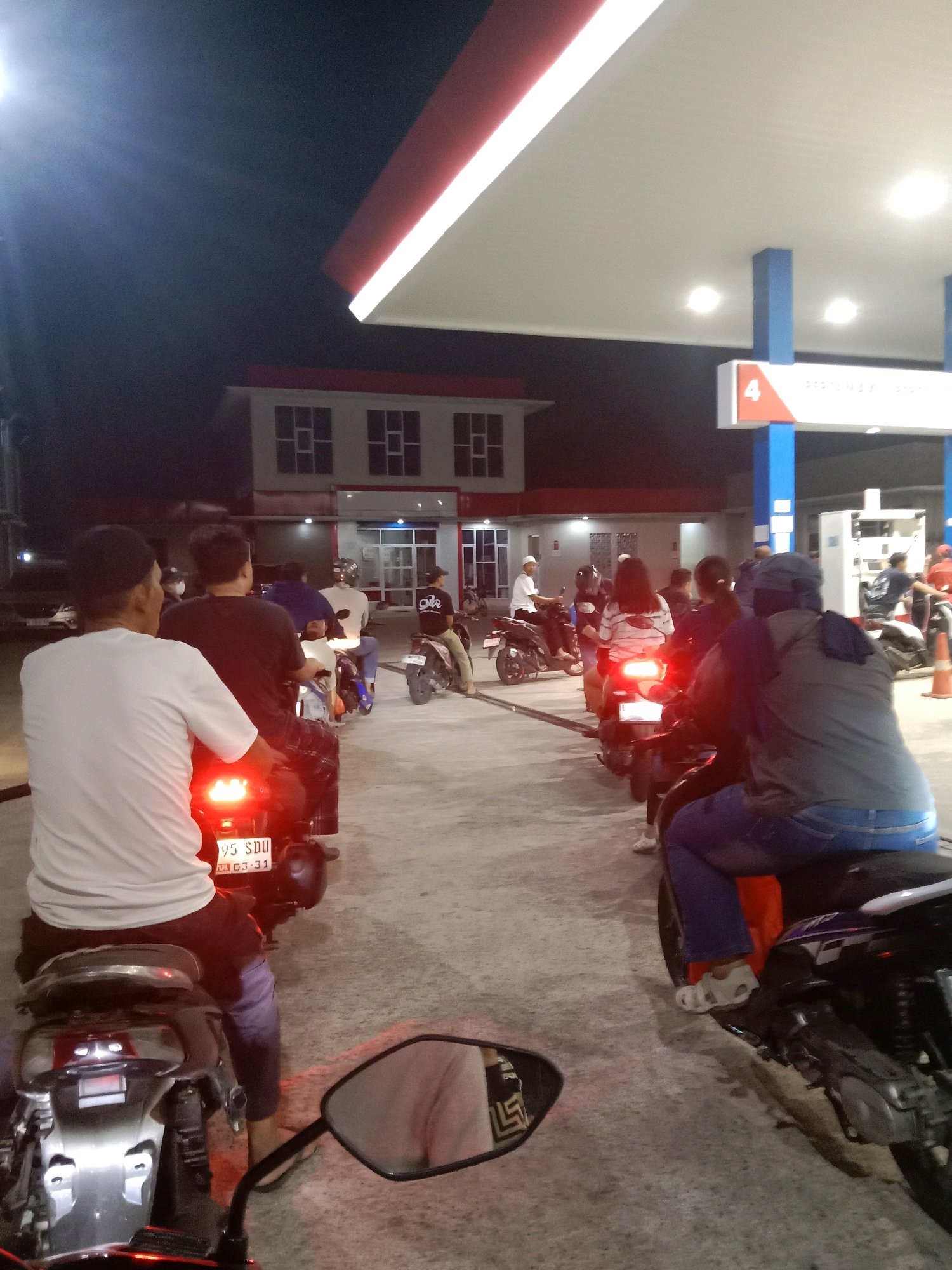 The image depicts a nighttime scene at a gas station, likely a Pertamina station in South Tangerang, Indonesia, given the presence of the Pertamina branding. Several people are on motorcycles, forming a queue. The foreground shows the rear of a motorcycle with its brake lights illuminated. Behind it, a person in a white t-shirt is seated on a motorcycle. Further back, a line of motorcycles and riders extends towards the gas pumps. The gas station canopy with its distinctive red and white trim is visible, as are the gas pumps and the station building. The lighting suggests it is dark outside, with artificial lights from the gas station illuminating the scene. The overall atmosphere is one of a typical evening at a busy public service location.