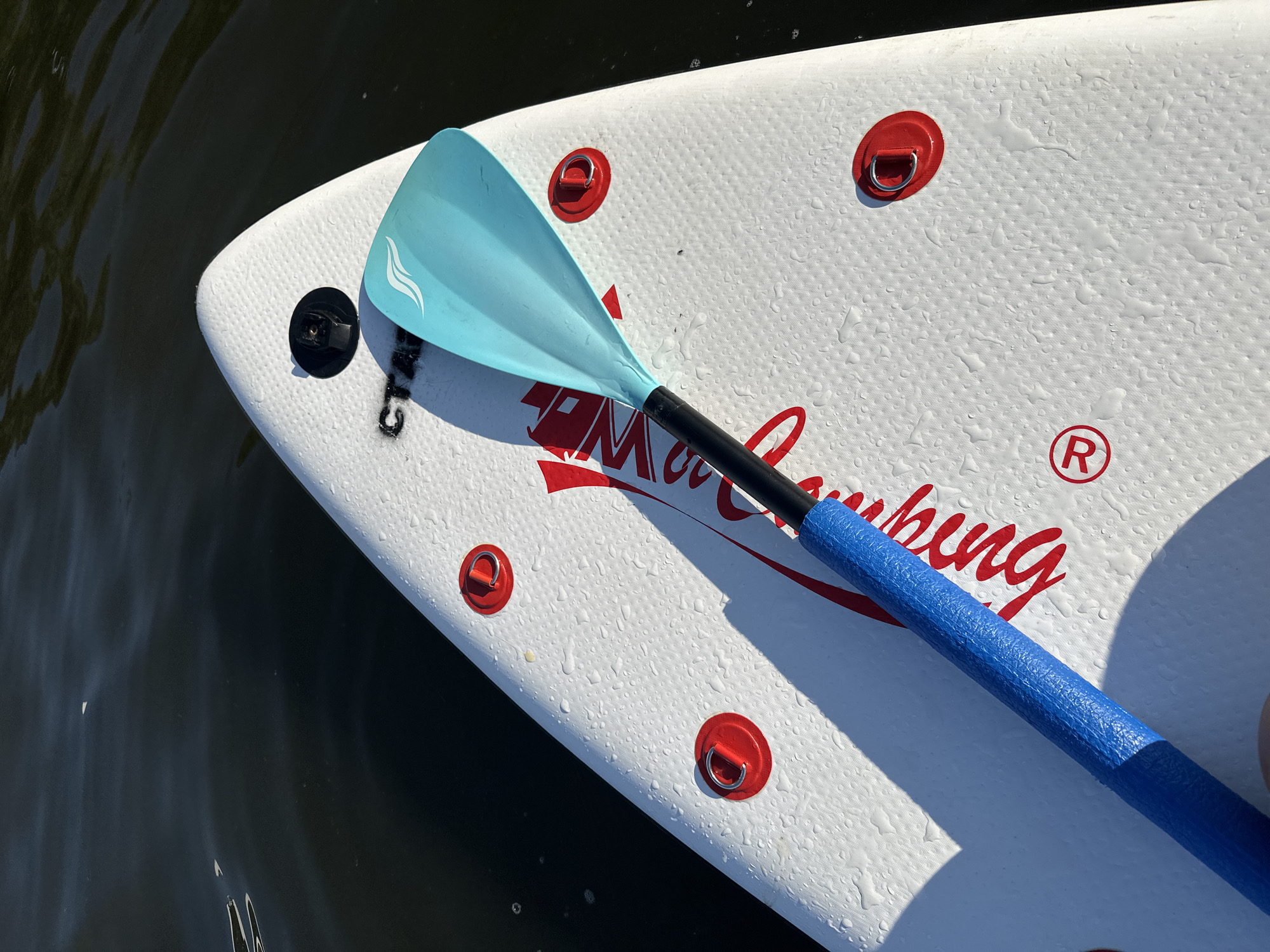 The image shows a close-up, top-down view of a white paddleboard floating on dark water. A blue and black paddle rests diagonally across the board, with its blue grip towards the bottom left and the light blue blade positioned towards the top. The paddleboard is wet, with numerous water droplets visible on its surface. Several red D-rings are attached to the board, and red lettering with a circular registered trademark symbol is also visible. The environment appears to be an outdoor body of water, consistent with the location of Туристический Пансионат «Клязьминское водохранилище», Russia, likely during daylight hours given the shadows and bright reflections on the water. No people are visible in the frame.