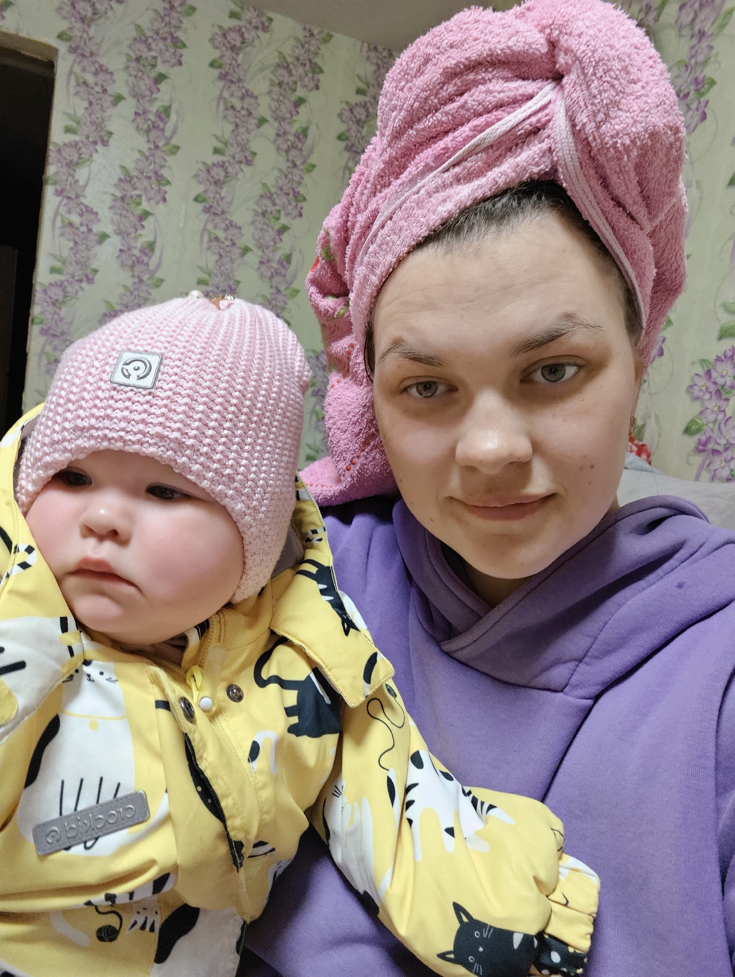 A woman is holding a baby. The woman has a pink towel wrapped around her head and is wearing a purple hoodie. The baby is wearing a pink knit hat and a yellow jacket with black and white cat patterns. The background shows a wall with floral wallpaper. The location is Уральское, Russia.