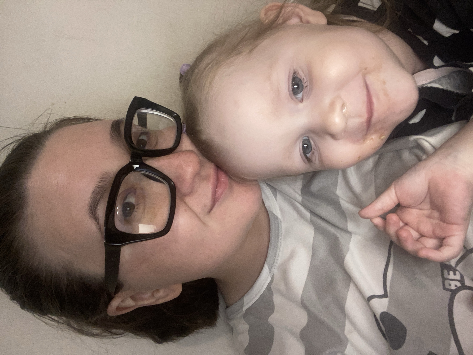 This media file captures an intimate, close-up portrait of an adult woman and a young child, likely a mother and daughter, lying down together indoors. The image is taken from a slightly elevated perspective, with the subjects oriented horizontally within the vertical frame.
The adult, on the left, has dark hair and is wearing large black-framed glasses. She is dressed in a light-colored top, possibly with grey and white stripes. Her expression is relaxed, with a subtle smile, and she looks directly towards the camera.
The child, a young girl with light blonde or light brown hair, is nestled closely beside the adult, with her head resting near the adult's temple. Her hair is partially gathered with a small purple elastic. She has bright blue eyes and a wide, joyful smile, also looking directly at the camera. Visible food smudges around her mouth and chin suggest she may have recently eaten. She wears a dark top with light patterns, possibly white stripes or polka dots. Her left hand rests on a grey and white patterned blanket or pillow, where partial text "BE" is visible.
Both individuals appear happy, comfortable, and affectionate, conveying a warm and tender mood. The setting suggests a cozy indoor environment, possibly a bed or couch, with soft, even lighting. The location context is Chaikovsky, Russia.