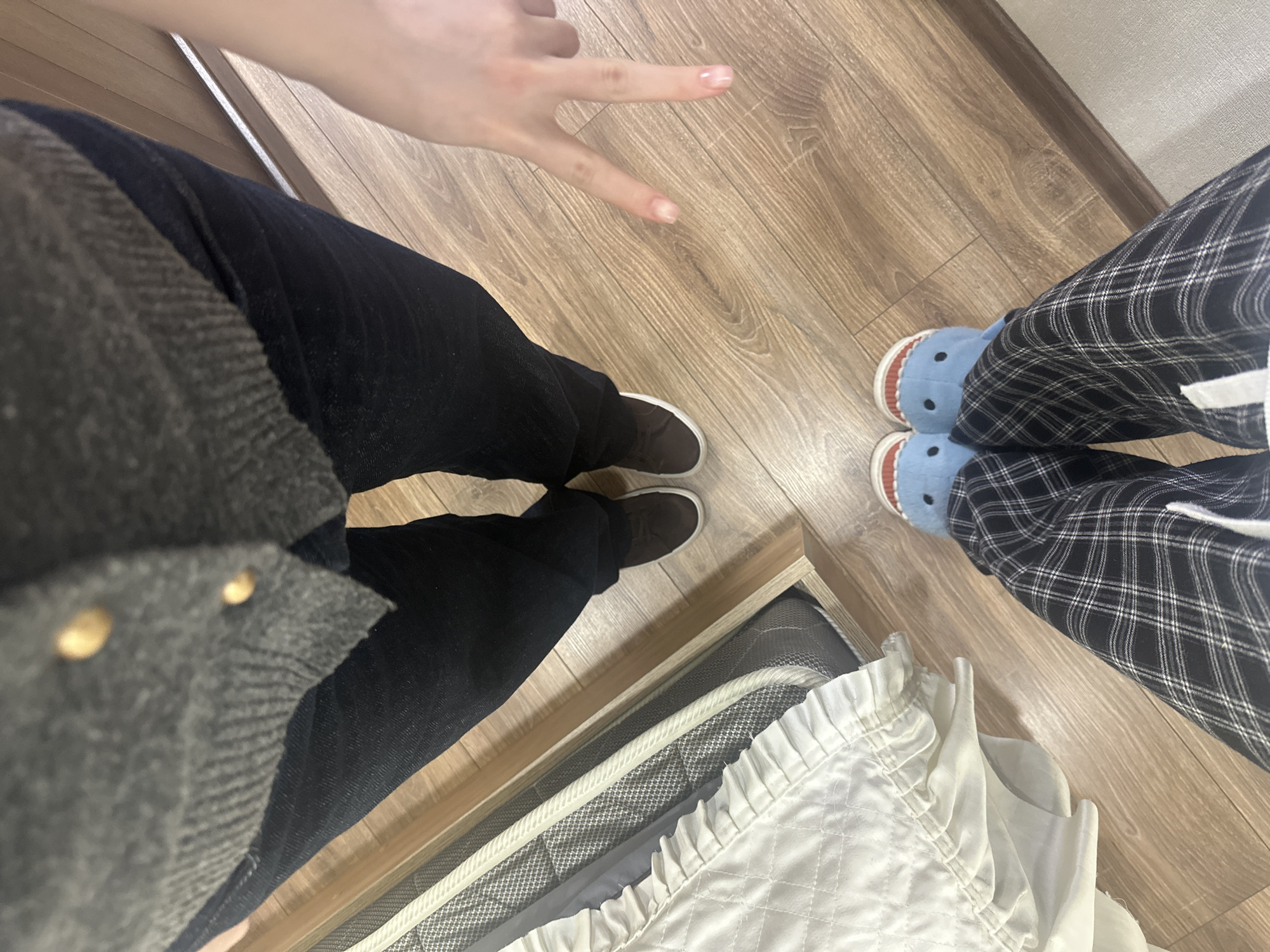The image presents a floor-level view of three sets of legs and feet, along with a section of what appears to be a bed. The person on the left wears dark jeans and brown shoes with a gray jacket or top. A hand makes a peace sign above the left set of legs. The person on the right is wearing plaid pants and blue, shark-shaped slippers. The background is a wooden floor. A white bedspread and a bed frame are near the bottom of the image. There is no information available in this image to determine the location of this scene as Yerevan, Armenia.