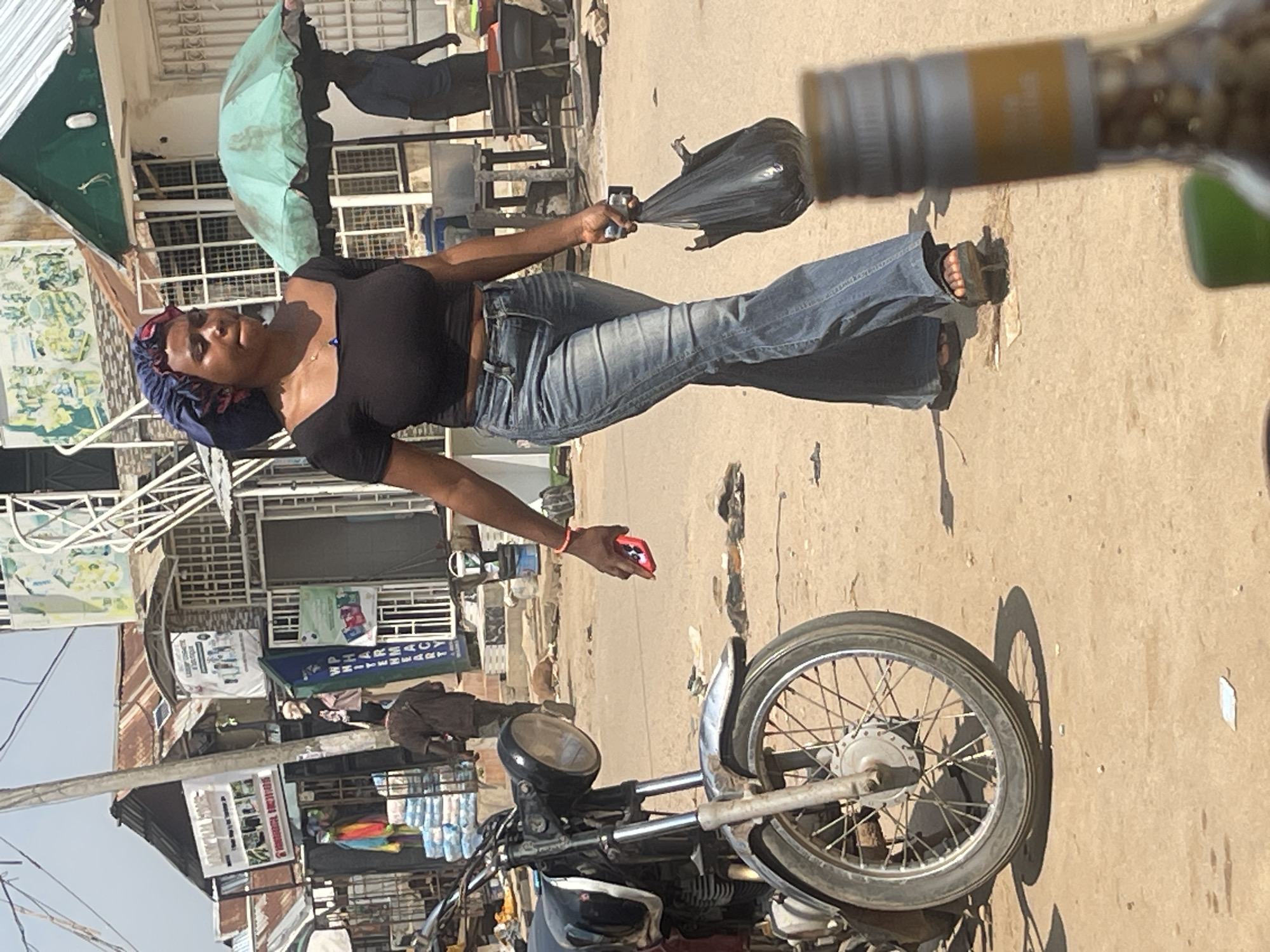 In an outdoor market scene in Abuja, Nigeria, a young Black woman walks from left to right, carrying a black plastic bag in her right hand and a red mobile phone in her left. She is wearing a black crop top, blue flared jeans, and dark sandals. A blue bonnet covers her hair. In the foreground, to the left, is the front half of a motorcycle. Behind the woman, to the right, a green umbrella shades a stall, where a person can be seen standing. In the background, various market stalls and buildings with corrugated metal roofs are visible, along with a power pole and hanging signs. The ground is unpaved and dusty.