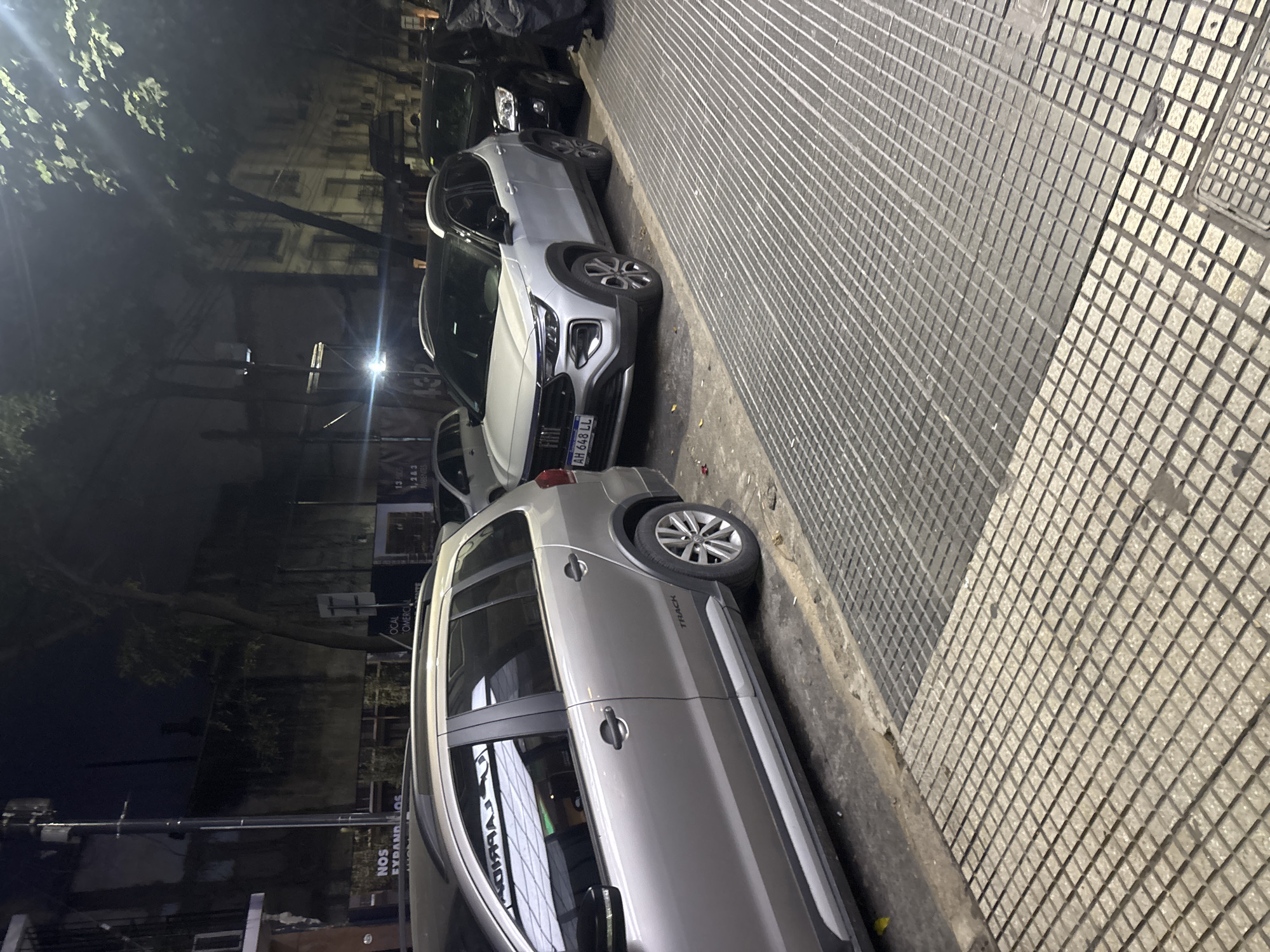 The photograph depicts a street scene in Buenos Aires, Argentina, at night. The camera's perspective is from ground level, looking towards the curb. Two parked cars are visible; a light-colored Volkswagen Saveiro Track parked closer to the camera and a light-colored Fiat Pulse further away. A dark-colored vehicle is parked behind the Fiat Pulse. The Fiat Pulse license plate is partially obscured but appears to read "AH 648 LL." The road surface is paved and the sidewalk is covered in tiles, with some variation in texture. A street light illuminates the background. Signs and building facades are visible in the background, suggesting a commercial or residential area. No people are visible in the frame.