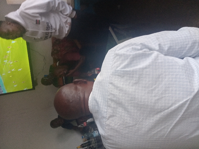 A gathering of people is watching television. A bald man in a light blue patterned shirt is in the foreground, his back to the viewer. Another man wearing a white sweatshirt is looking at the television screen. The television screen shows a green background and appears to have graphics resembling a sporting event. Water bottles and other items are on the floor.
