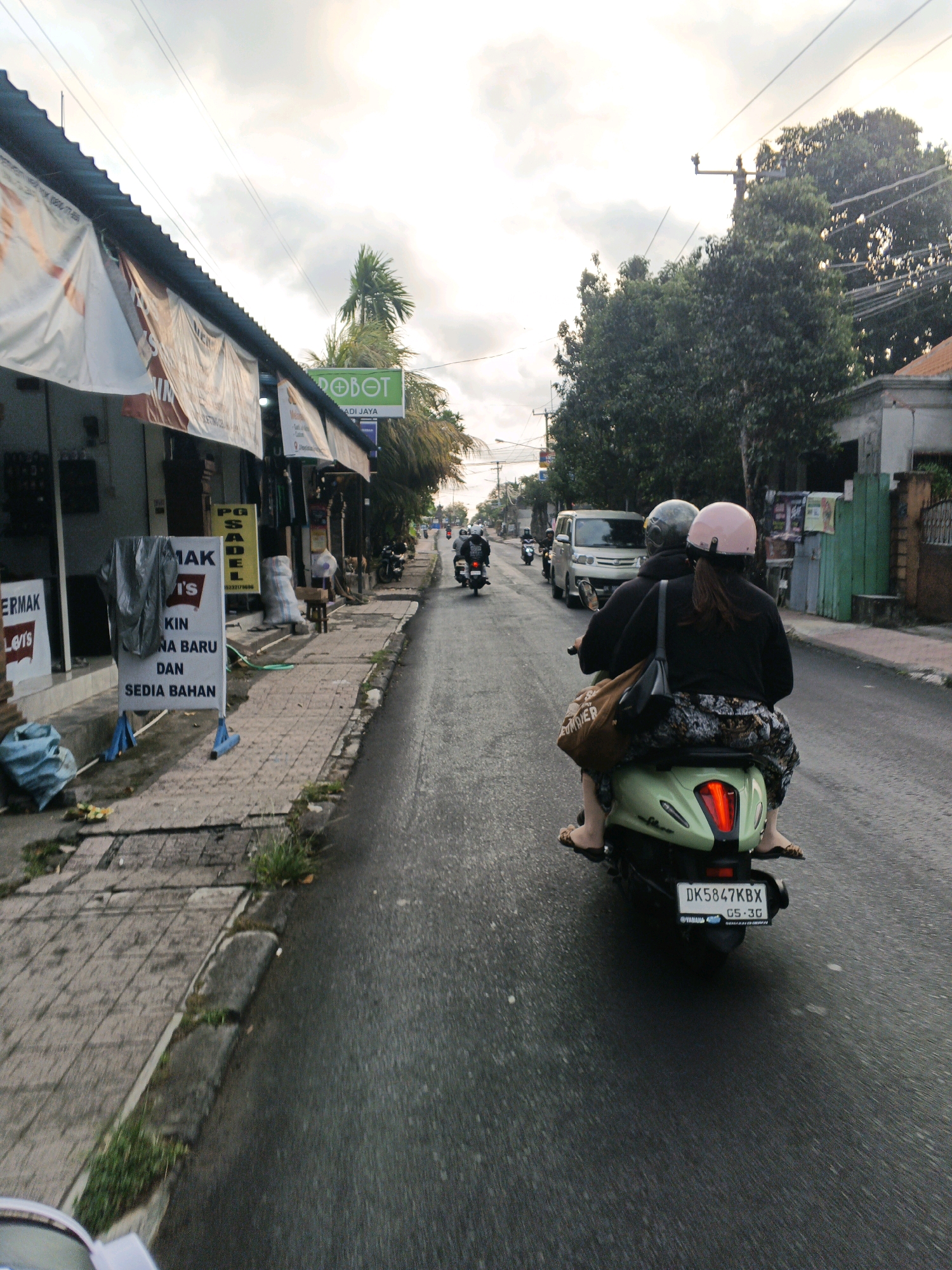 This is a street-level, horizontal, full-color, medium-shot photograph taken from the perspective of someone riding on the back of a scooter. The scene is set in Sukawati, Indonesia. In the foreground, the back of a light green scooter is prominent, with a license plate reading "DK5847KBX" and "GS-3G". Two people are seated on the scooter, both wearing helmets, one a dark grey and the other a light pink. The passenger in the back is wearing a patterned skirt and flip-flops. A brown tote bag is slung over the passenger's shoulder. The scooter is moving on a wet asphalt road, suggesting recent rain.
To the left of the road, a sidewalk is paved with brick tiles and bordered by a low concrete curb. Several shops line the sidewalk, with corrugated metal roofs. Banners are hung from the shop fronts, one displaying the word "ROBOT" in green and white. Signs in Indonesian are visible on the shop doors and on a freestanding sign on the sidewalk, which reads "MAKMUR JAYA" and displays text related to clothing. Blue plastic bags and other debris are scattered on the sidewalk.
In the middle ground, other vehicles are visible on the road, including a silver van and several motorcycles, all moving in the same direction as the photographer. Trees and buildings can be seen on the right side of the road, with power lines crisscrossing overhead. The sky is overcast, with diffused light suggesting either early morning or late afternoon. The overall atmosphere is that of a typical urban street scene in a tropical region.