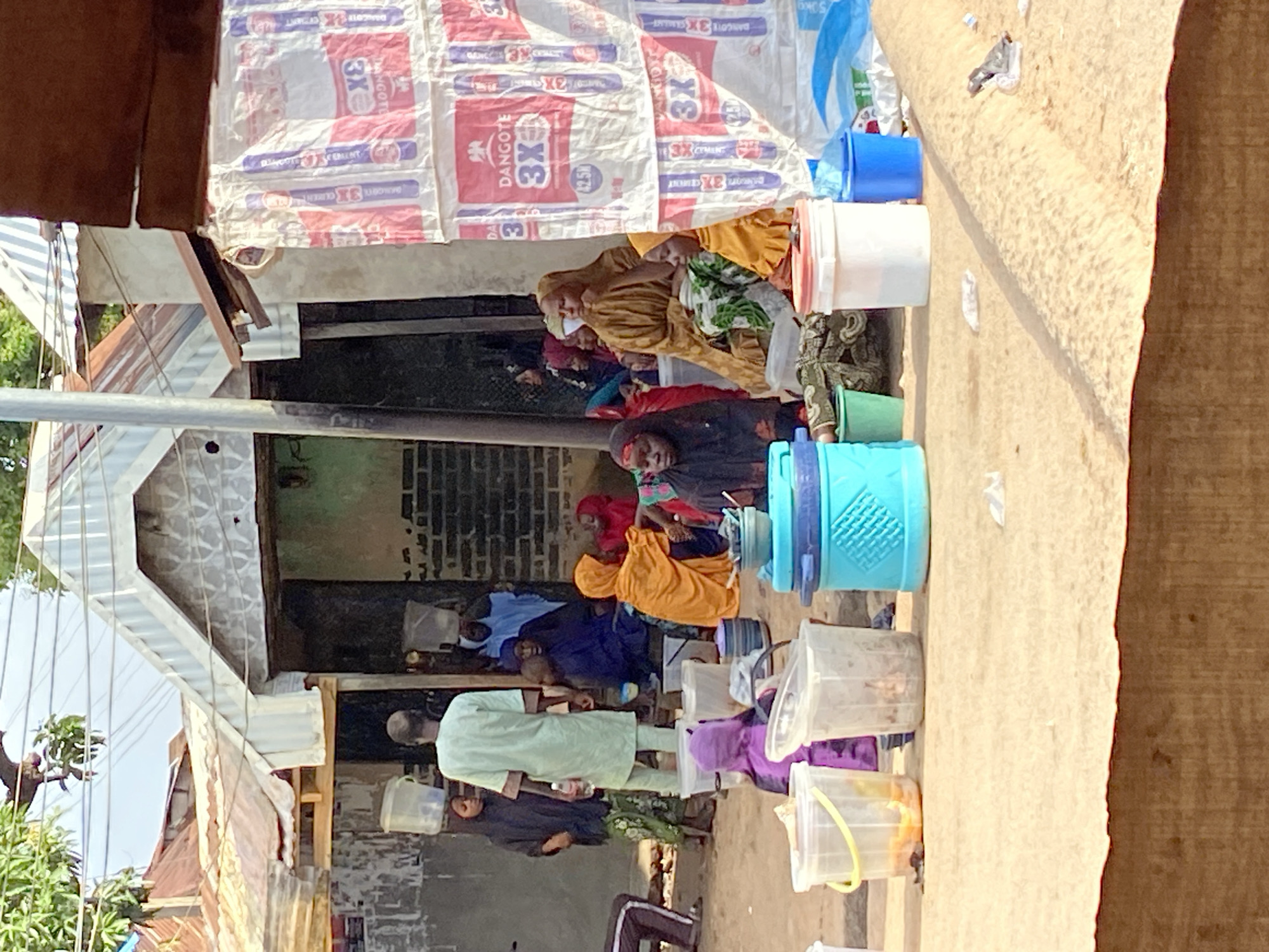 In Mararaba, Nigeria, a community gathers outside a building with corrugated metal roofs. A man in a light green garment stands near the entrance, holding a water bottle. Several individuals, mostly women and children, are seated or standing around him, many wearing colorful headscarves and traditional clothing. Some are carrying buckets on their heads or holding them at their sides. In the foreground, various plastic containers, including large buckets and smaller tubs, are placed on the ground. The scene suggests a communal activity, possibly related to water collection or distribution. The environment appears to be an outdoor marketplace or residential area, with buildings constructed from various materials.