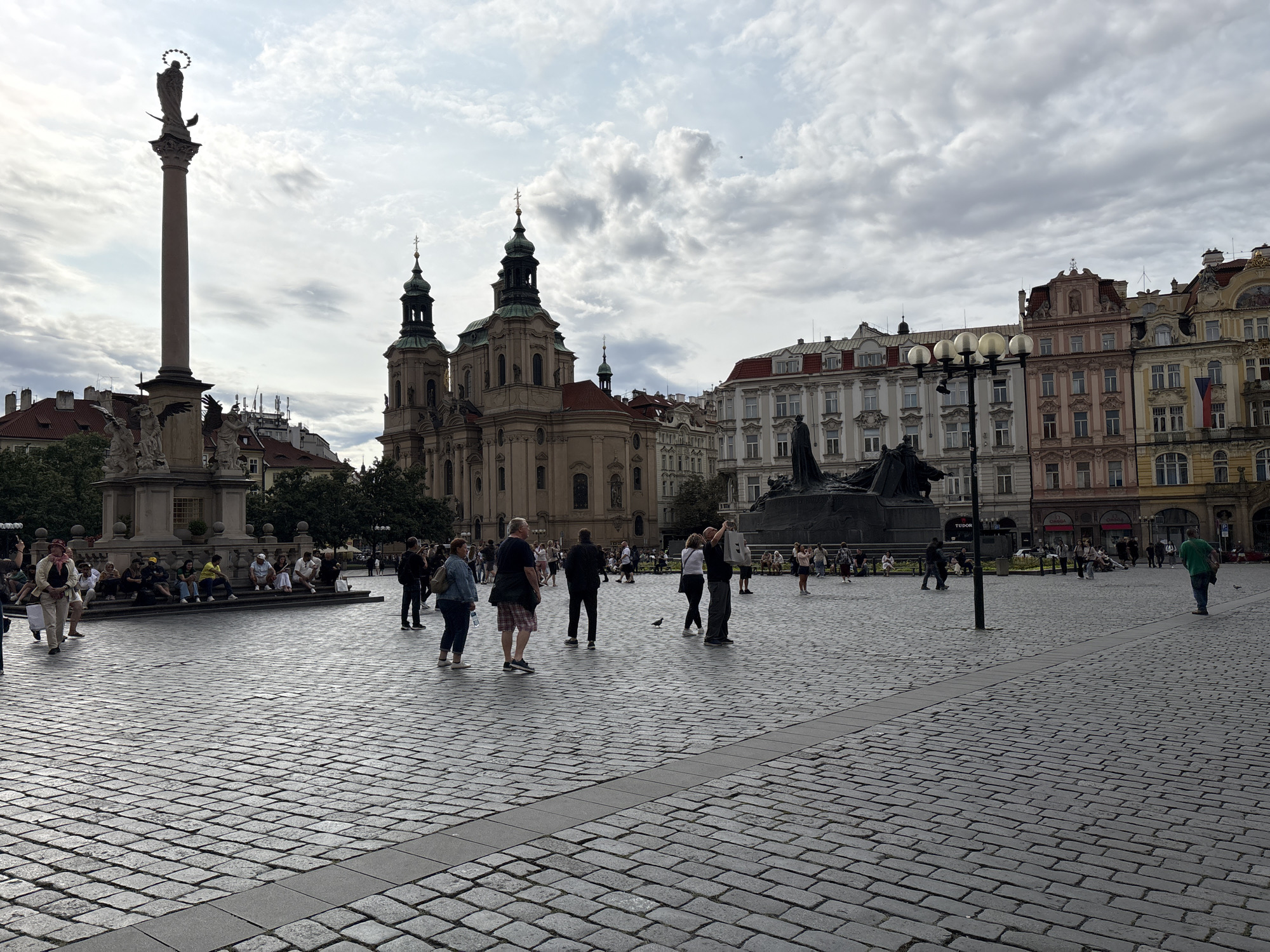 This is a medium-wide shot of the Old Town Square in Prague, Czechia. The square is paved with cobblestones and several people are present, some walking and some sitting. In the background, a large, ornate church with two spires dominates the left side of the frame. To the right of the church are several multi-story buildings with varied architectural styles and colored facades. In the foreground, on the left, is a tall column topped with a statue. A large monument with a statue on horseback is visible in the mid-ground to the right. The sky is cloudy.
