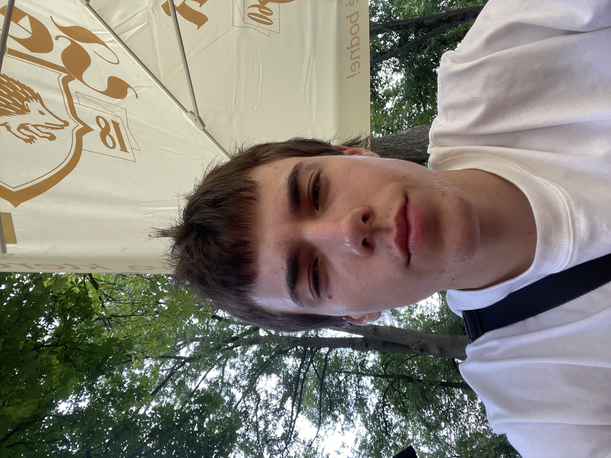 A young, light-skinned male with brown hair and light brown eyes is pictured in a close-up, front-facing selfie. He is wearing a white t-shirt and has a black strap across his left shoulder. The background consists of green trees and a white umbrella with orange lettering. The scene appears to be outdoors, possibly in Jihlava, Czechia, given the umbrella's design which resembles those commonly found at outdoor seating areas of establishments like beer gardens or cafes. The lighting suggests it is daytime.