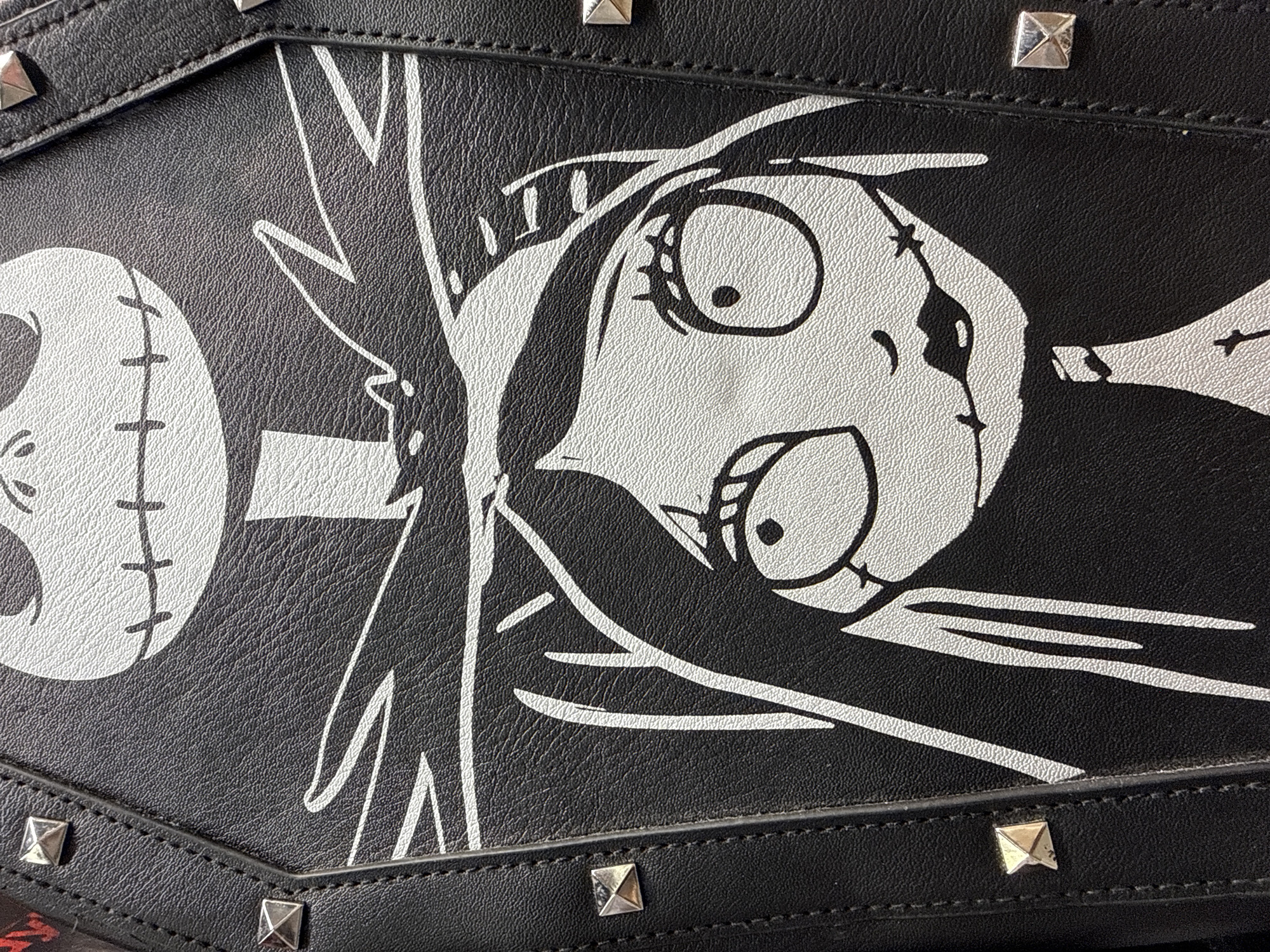 The image is a close-up, high-angle, vertical shot of a black coffin-shaped bag with a white graphic of Sally from "The Nightmare Before Christmas." The bag is made of textured faux leather and is adorned with silver pyramid studs along its edges. The graphic features Sally's face prominently, with her characteristic large, round eyes, stitched mouth, and long, dark hair. Above Sally's head, the top portion of Jack Skellington's skull face is visible, also in white. The image is taken indoors, and there are no discernible environmental details that would place it in Madrid, Spain.