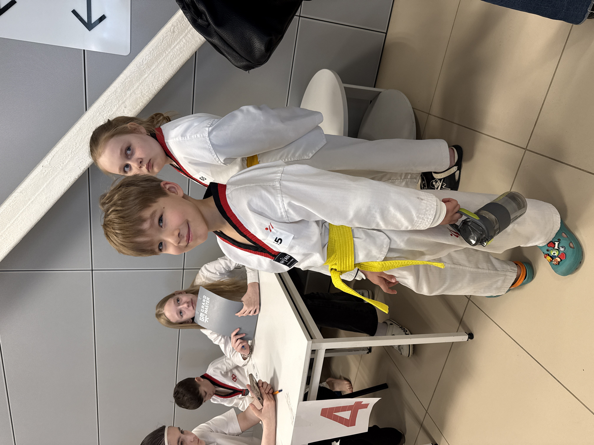 A group of children in white martial arts uniforms with colored belts are standing around a white table in an indoor area, seemingly a waiting or registration area. One child is holding a water bottle. There is a sign with a downward-pointing arrow on the wall. A folder with text “GM GRAND MASTER” is visible at the table. The location is presumed to be a building in the city of Rostov-on-Don, Russia.