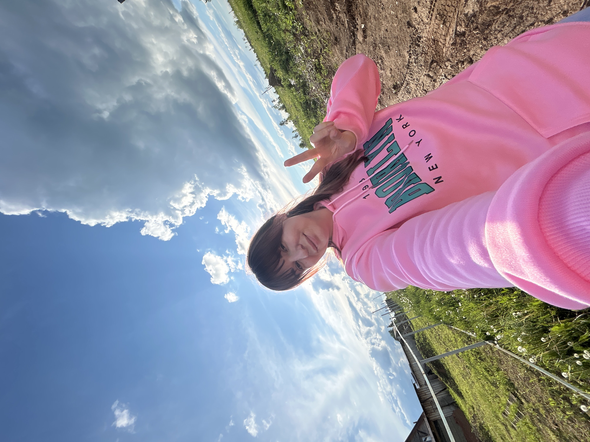 A woman is holding up a peace sign while standing outside in Фоки, Russia. She is wearing a pink hoodie with "Brooklyn New York 1983" printed on it. The sky is blue with scattered clouds. There is green grass and some unidentified structures visible in the background.