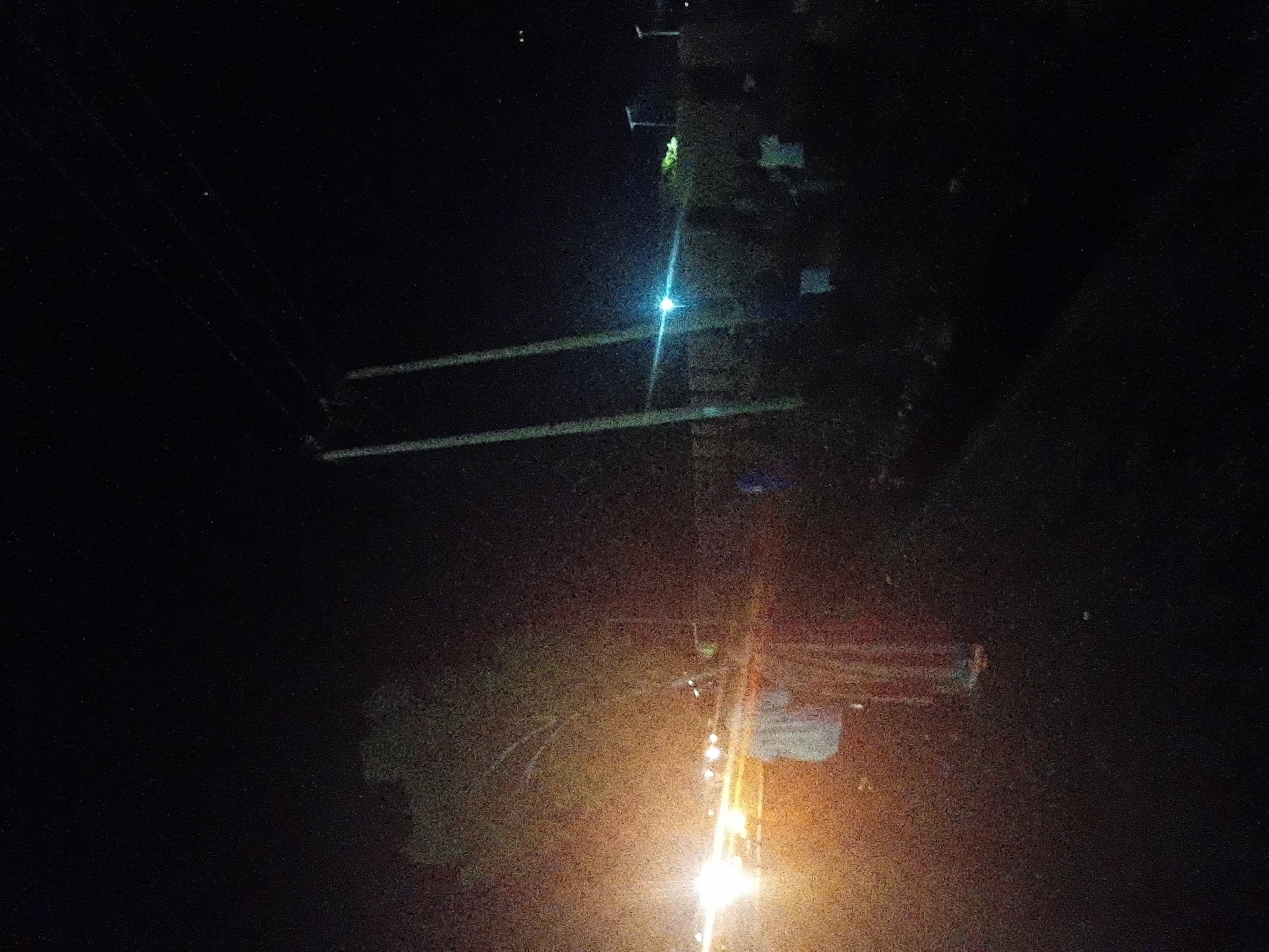The image depicts a nighttime roadside or street scene in Jalingo, Nigeria, characterized by significant image noise and varied artificial lighting. On the far left, an intense, diffused white and yellow light source, consistent with vehicle headlights, illuminates the immediate foreground and mid-ground. Within this illuminated area, a single individual is visible, standing or walking, clad in a light-colored top and dark lower garments. Behind this individual, a very indistinct, reddish form is discernible.
Towards the mid-right, two prominent, slender utility poles, likely concrete or wood, stand vertically, with faint overhead lines extending diagonally. A distinct blue-white light source, consistent with a streetlight, is situated in the mid-ground, slightly to the right of these poles. Several indistinct human figures are visible beneath and around these utility structures, some wearing light-colored tops. The background beyond the illuminated areas is predominantly dark, with faint suggestions of foliage and additional distant pole-mounted lights visible towards the far right horizon. The ground appears dark, with some light reflection in the headlight-illuminated section.
