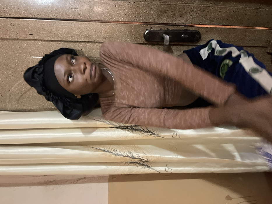 A young adult female stands indoors in Muchiya, Nigeria, positioned between a light-colored curtain and a wooden door. She has a dark complexion and wears a black satin bonnet, a light brown long-sleeved crop top, and blue shorts featuring white and green patterns. A thin, silver-colored necklace is visible on her neck. Her lips are pursed, and her gaze is directed towards the viewer's right. Her arms are blurred, indicative of motion, with one arm positioned across her lower abdomen. The background on the left features a cream or beige curtain adorned with dark, feather-like patterns, while on the right, a textured wooden door with a dark metal handle and keyhole assembly is visible. A plain light brown wall is partially visible behind the curtain.