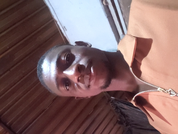 A dark-skinned man with closely cropped hair poses for a selfie indoors. The camera angle is low, capturing his face and the upper part of his brown collared shirt with a visible zipper. The lighting is warm, casting shadows on the left side of his face. A wooden wall is visible in the background, contrasting with a brighter, lighter colored wall or surface above it.