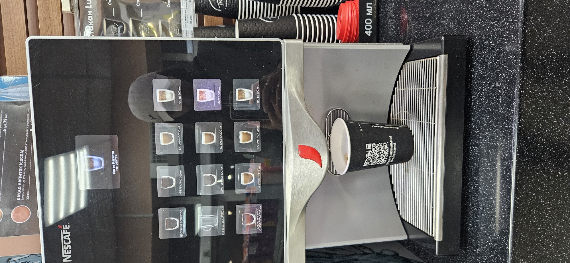 The image depicts a Nescafe coffee machine located in a retail setting in Vitsebsk, Belarus. The machine has a digital display showing various coffee options, with an active selection highlighted as "Латте Накиято готовится" (Latte is preparing). A black paper cup with a QR code and text in Russian is placed on the drip tray, filled with a coffee beverage. To the right of the machine, stacks of black and white disposable cups are visible, along with a red lid and a sign indicating "400 мл". The background suggests an indoor commercial space, possibly a convenience store or a cafe counter. A person wearing a dark cap and hoodie is partially visible, reflected in the machine's screen, obscuring some of the background details. The overall scene is a straightforward depiction of a coffee vending machine in operation.
