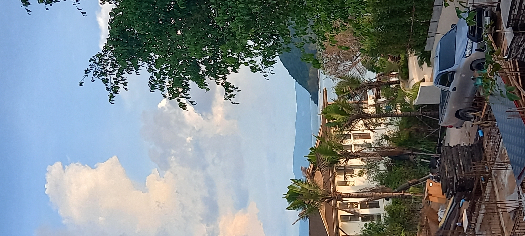 Baan Wok Tum, Thailand. A daytime, outdoor scene capturing a partial view of a building adorned with palm trees, observed from an elevated perspective, likely a construction site. To the right, a light-colored pickup truck parked adjacent to building materials. In the background, a mountain and sea visible. A mix of clouds and sky are visible to the left, partially obscured by foliage.