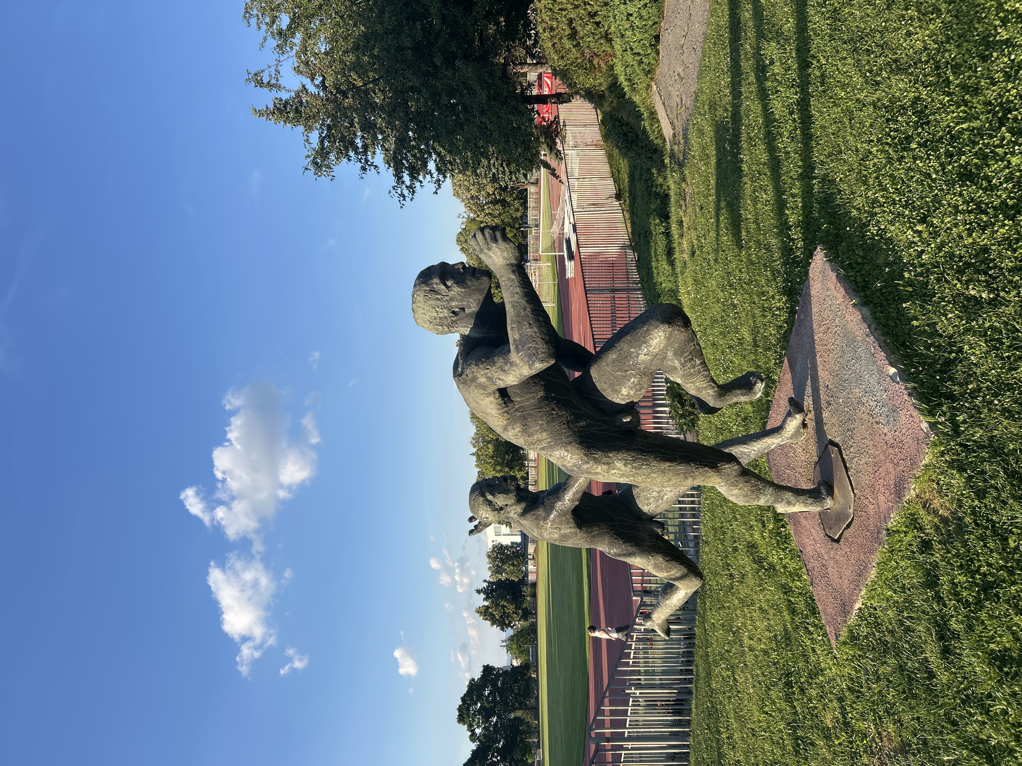 The image shows a bronze sculpture of two figures in a dynamic pose, set outdoors on a grassy area with a running track and stadium seating in the background. The sculpture appears to depict a scene of struggle or intense physical exertion, with one figure appearing to be pushing or pulling the other. The figures are rendered in a realistic style, with muscular anatomy and expressive faces. The sculpture is placed on a reddish-brown stone base.
The scene is set under a clear blue sky with scattered white clouds, suggesting a pleasant day. The sun is casting long shadows from the sculpture and surrounding trees, indicating it is likely late afternoon or early evening. The lush green grass and vibrant foliage suggest a warm season, possibly spring or summer. In the background, a running track with red and green lanes is visible, along with white bleachers of a stadium. Beyond the stadium, more trees and a building can be seen. The overall setting appears to be a public park or sports complex.
The image is captured from a low angle, looking up towards the sculpture and the sky. The orientation of the photo is such that the sky is on the left and the ground is on the right, suggesting the photographer may have been standing on a slope or the image has been rotated. The sculpture itself is the central focus, with the background providing context for its placement. The mood of the image is one of outdoor activity and public art, with the bright sky and natural surroundings contributing to a sense of openness and vitality. There is no visible text in the image.