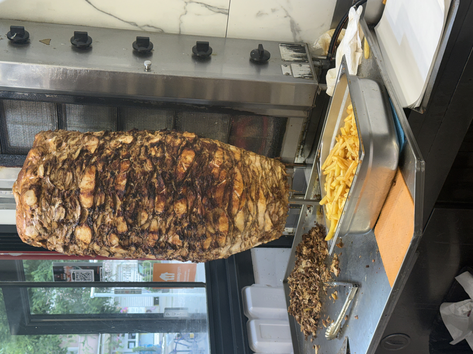 A vertical spit of doner meat, browned and layered, is positioned under a stainless steel grill in a restaurant setting. To the right, a metal tray is filled with french fries. Below the tray, there is a pile of shaved meat and a pair of tongs. A window is visible in the background, reflecting an outdoor scene, suggesting the restaurant is located near a street with foliage visible. The setting is likely a commercial kitchen environment, possibly in a fast-food restaurant. The exact location in Plovdiv, Bulgaria, cannot be determined from the image alone.