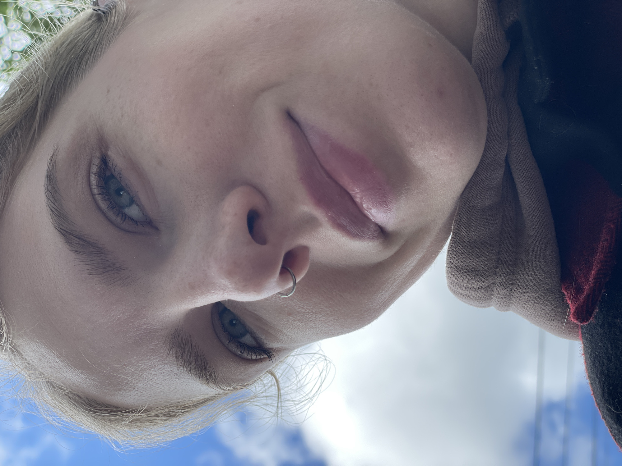 A close-up, slightly skewed perspective photograph features a person's face. The person has light skin, blue eyes, and blonde hair. A silver ring is visible in their left nostril. Part of a garment is seen on the right side of the frame, with colors ranging from tan to red and dark gray. The background shows a glimpse of blue sky with white clouds. The location is not discernible as Izhevsk, Russia.