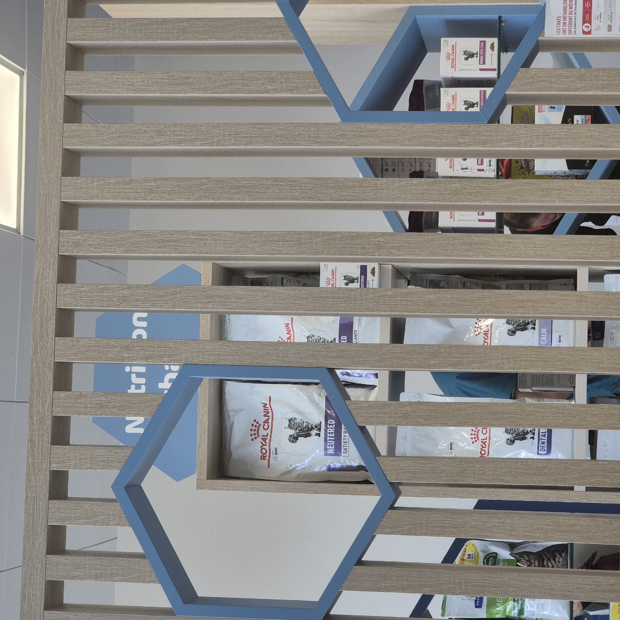 The image shows a section of shelving displaying various pet food products, likely cat food, primarily from the brand Royal Canin. There are also products from Hill's and Ntrion. The shelving unit is constructed from light brown, wood-look slats with a geometric blue trim. The environment appears to be a retail setting, potentially a pet supply store or veterinary clinic. The presence of French text ("LES CHATS ONT UN METABOLISH DIFFERENT DU NÔTRE") suggests the location is in a French-speaking area. The location within the city of Geispolsheim, France is not directly apparent from the image.