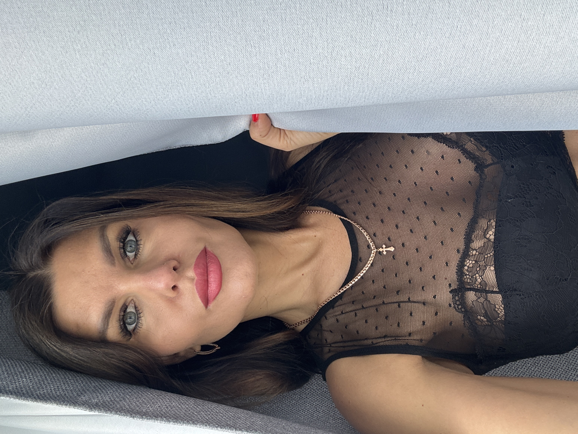 A woman lies on a gray surface, possibly a couch or bed, under a white sheet or fabric. Her head is resting on the surface, and she is looking at the camera. She has brown hair, blue eyes, and pink lips. She wears a black sheer top with small dots and a black lace bra underneath. A gold necklace with a cross pendant is visible around her neck, along with gold earrings. Her right hand, with red nail polish, is holding the edge of the white fabric above her. The location is not discernible from the image.