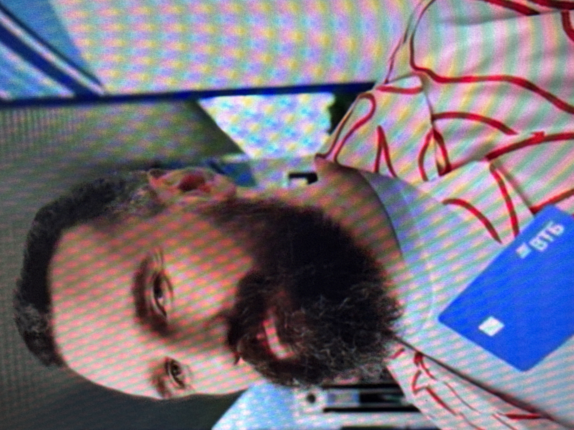 The image shows a close-up of a man with dark hair and a full beard, looking towards the left. He is wearing a light-colored shirt with a red, web-like pattern. In the foreground, a bright blue card with a white square and the letters "ВТБ" is visible. The background is abstract and appears to be a digital display with a pixelated pattern of various colors, suggesting an indoor setting possibly related to advertising or media. There is no identifiable information to place this image in the city of Saratov, Russia.