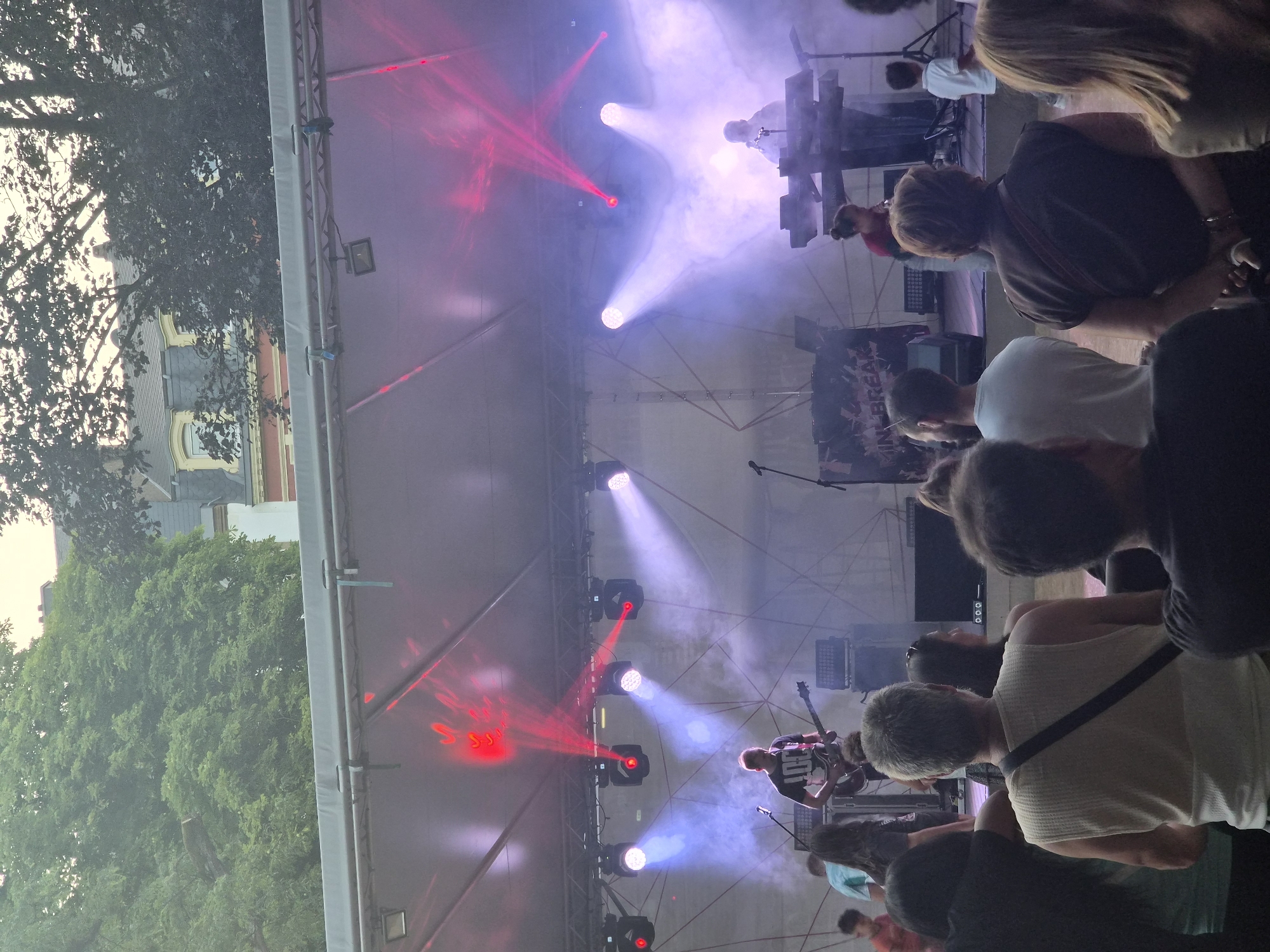 The image shows an outdoor concert or music festival taking place on a stage in Differdange, Luxembourg. A band is performing on stage with several band members visible. One person is playing a guitar, and another is playing a keyboard. The stage is lit with red and white lights, and there is some smoke or fog on stage. The audience is visible in the foreground, watching the performance. The background shows trees and buildings, suggesting an urban or park setting. The overall atmosphere appears to be lively and engaging.