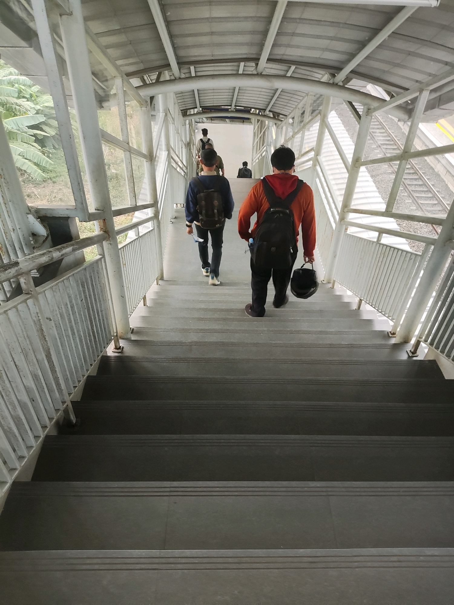 The image depicts a pedestrian overpass at a train station in Telagamurni, Indonesia. Four individuals are visible on the stairs of the overpass, all walking away from the camera. The person closest to the viewer, in the foreground, is wearing an orange hoodie and carrying a backpack, holding a black helmet in their right hand. To their left, another person in a dark blue jacket with a backpack is walking down the stairs. Two more individuals are further up the stairs, further away and less detailed. The overpass has a metal roof supported by white pillars and metal railings along the sides. To the right of the overpass, train tracks are visible. The environment appears to be somewhat overgrown with lush green vegetation, suggesting a natural setting surrounding the urban infrastructure.